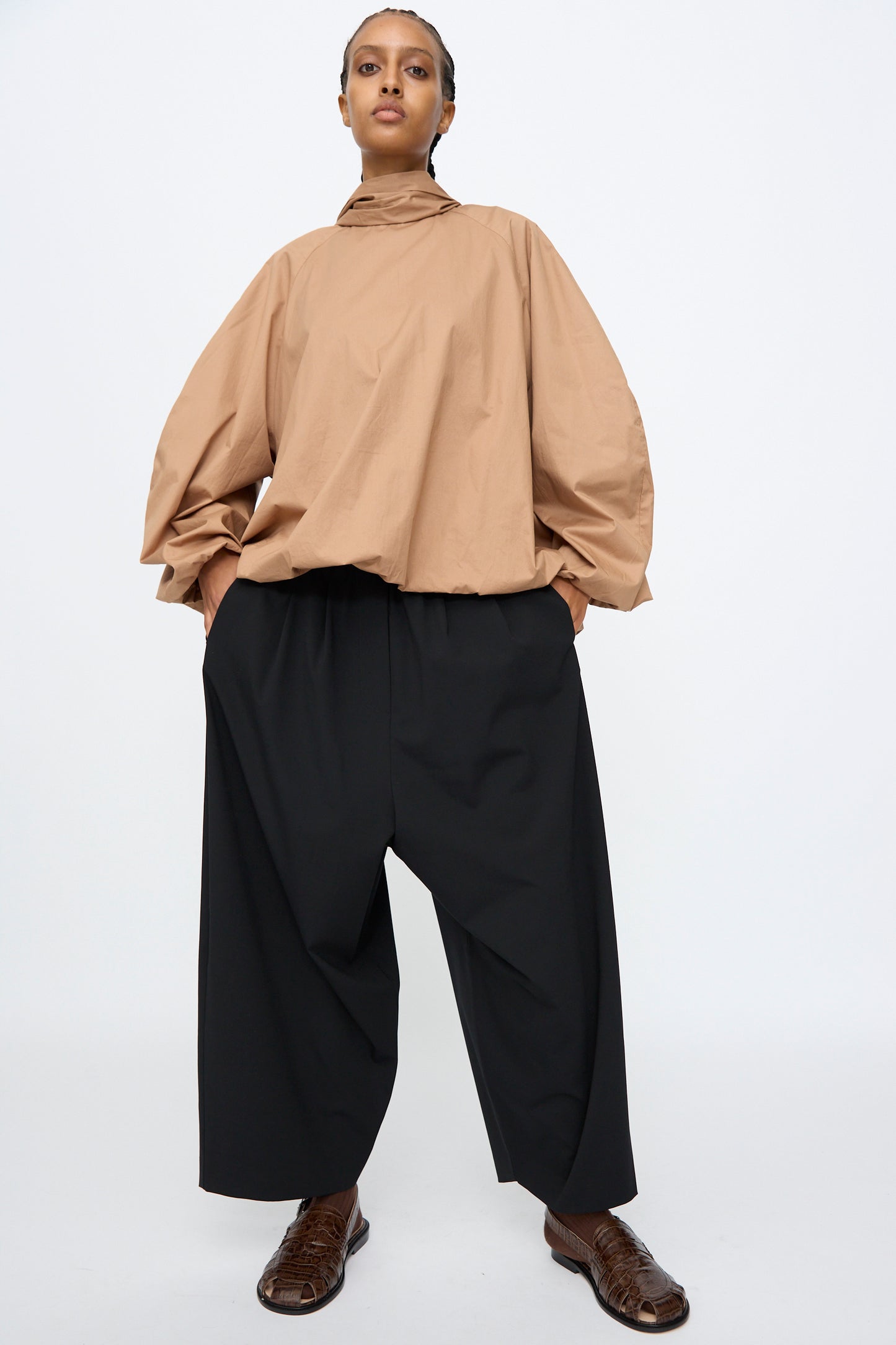 A person stands with hands in pockets, wearing a loose tan long-sleeve top and Cordera Tailoring Maxi Pants in Black with an elastic waistband, paired with brown shoes, against a plain white background.
