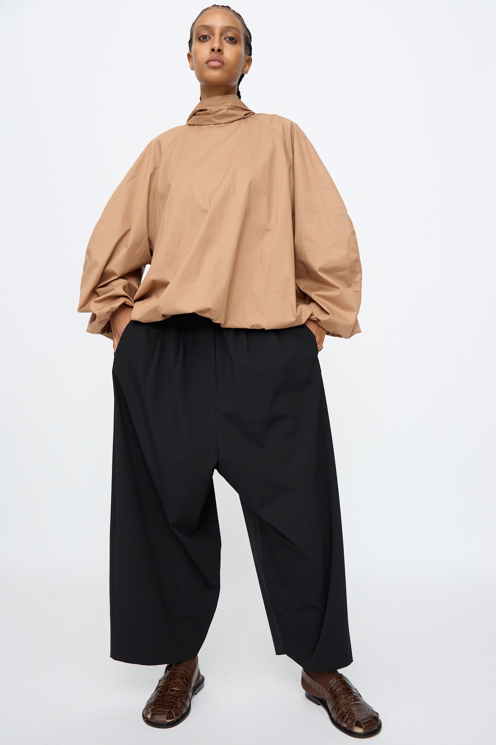 A person stands with hands in pockets, wearing a loose tan long-sleeve top and Cordera Tailoring Maxi Pants in Black with an elastic waistband, paired with brown shoes, against a plain white background.