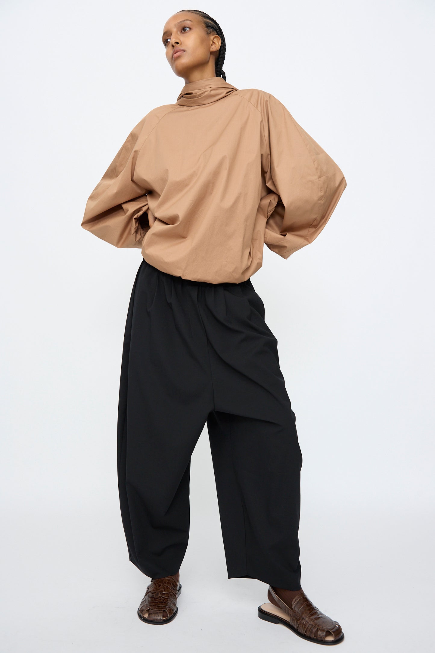 A person stands against a plain background wearing a loose tan top, Cordera Tailoring Maxi Pants in Black with an elastic waistband, and brown woven shoes.