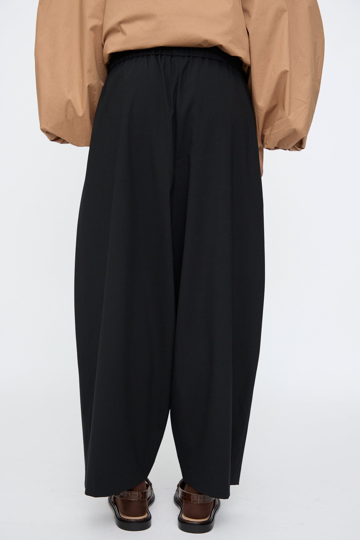 Model wears the Cordera Tailoring Maxi Pants in Black, featuring an elastic waistband, paired with a tan long-sleeve top and brown sandals, photographed from the back against a plain background.