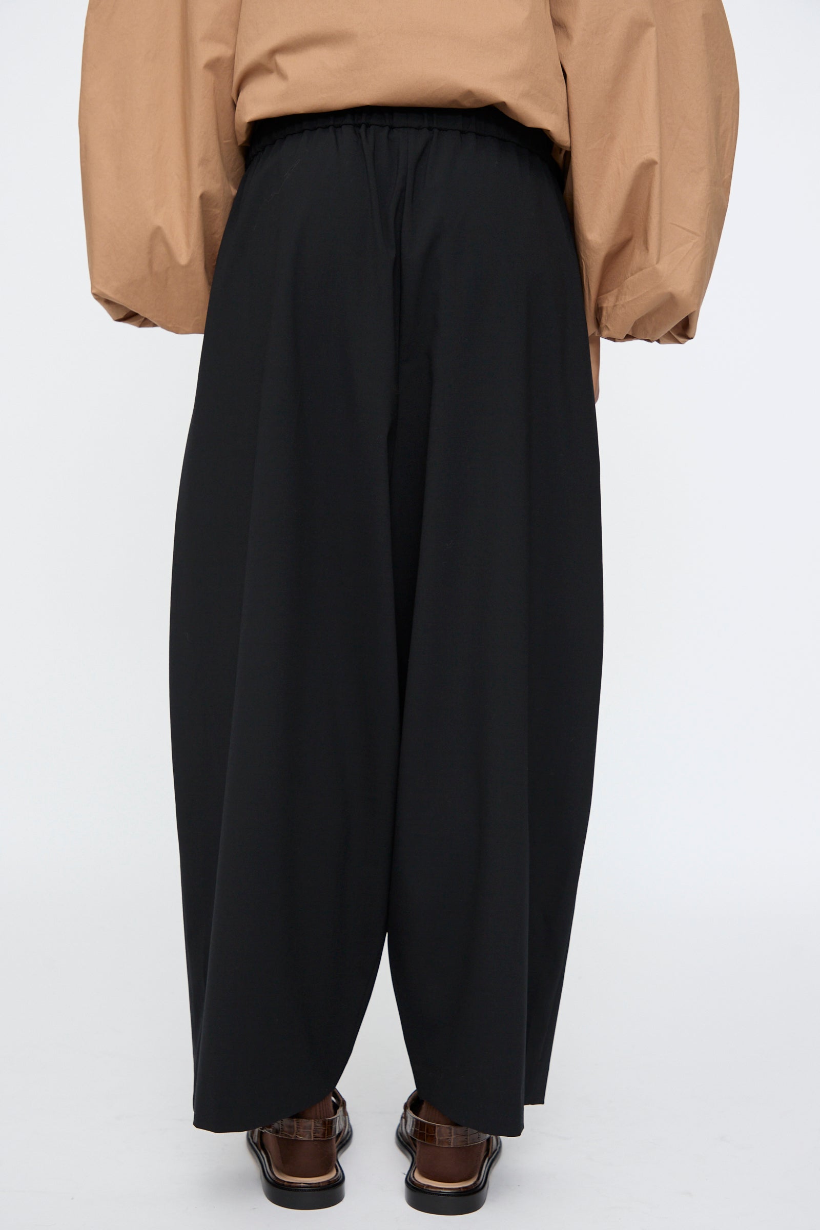 Model wears the Cordera Tailoring Maxi Pants in Black, featuring an elastic waistband, paired with a tan long-sleeve top and brown sandals, photographed from the back against a plain background.