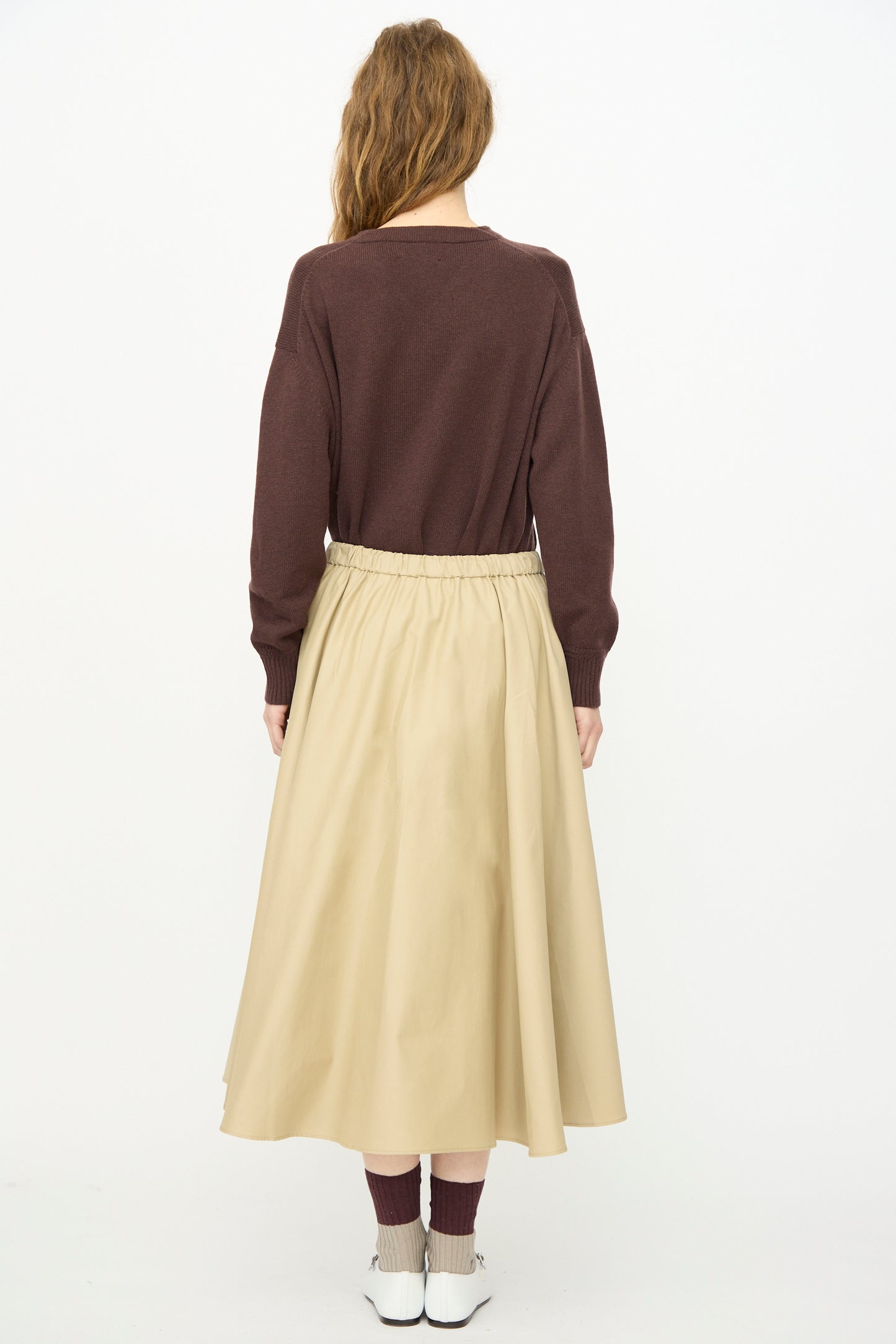 A person with long hair, wearing a brown sweater and Cordera's Technical Skirt in Camel, stands facing away against a plain white background, paired with brown socks and white shoes.