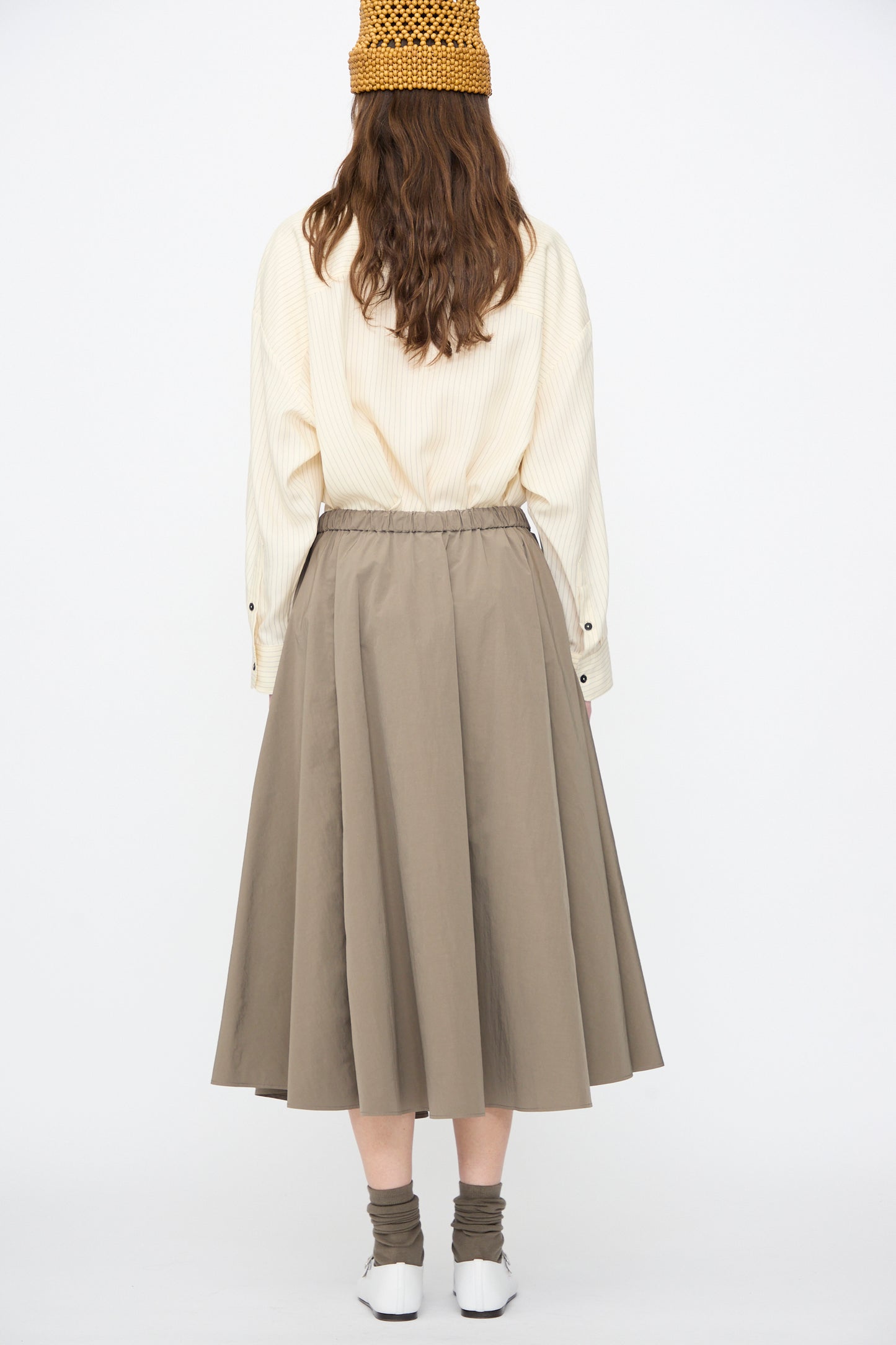 A person stands facing away, wearing a beige long-sleeve shirt, brown crochet hat, Cordera's Technical Skirt in Vetiver with an A-line silhouette and elastic waistband, white shoes, and brown socks.