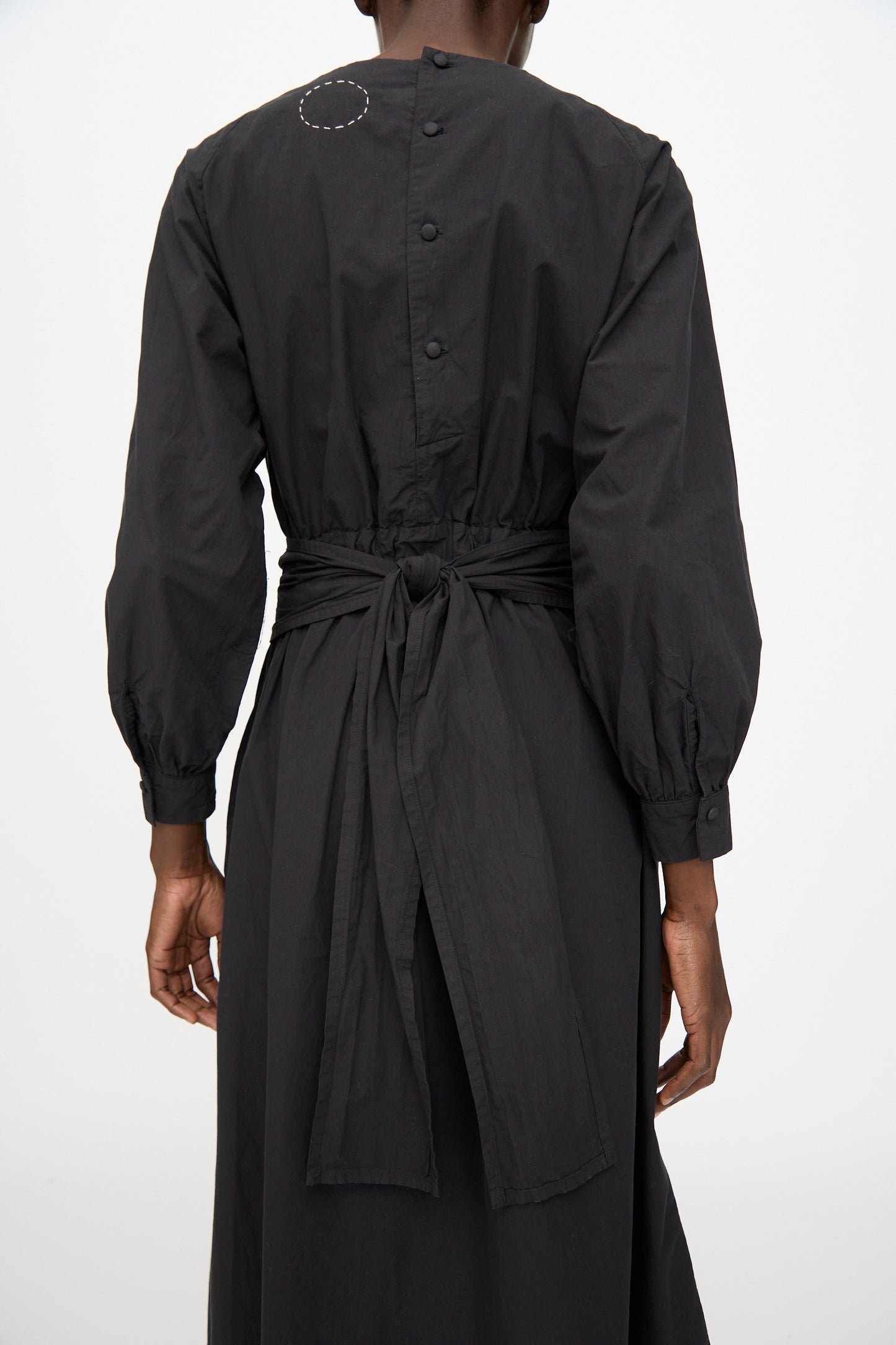 A person is seen from the back wearing the Cosmic Wonder Cotton Broadcloth Wrapped Dress in black, featuring long sleeves, a tied waist belt, and buttons along the upper back.