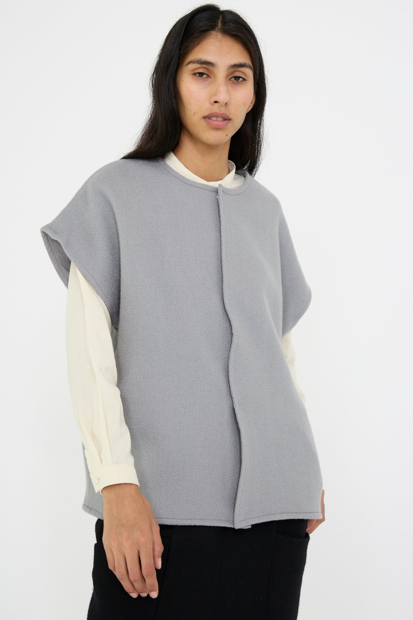 A person stands against a plain background, dressed in the Cosmic Wonder Herringbone Shetland Wool Dolman Sleeve Vest in gray, layered over a white shirt against a white background.