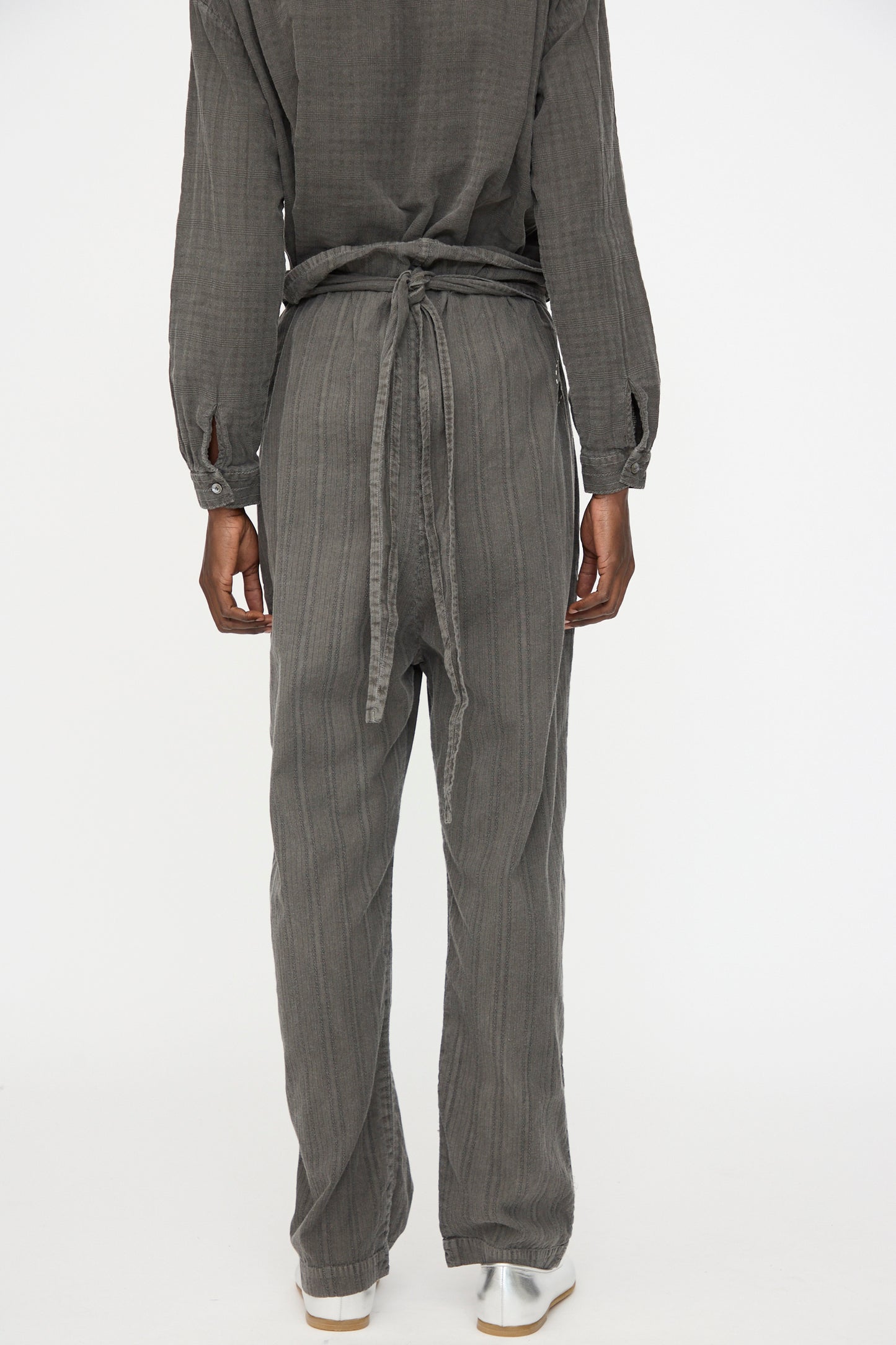 Person standing facing away, wearing Cosmic Wonder's Stripe Combination Wrapped Pant in Dark Sumi with a long-sleeved, gray textured top and white shoes, against a plain white background.