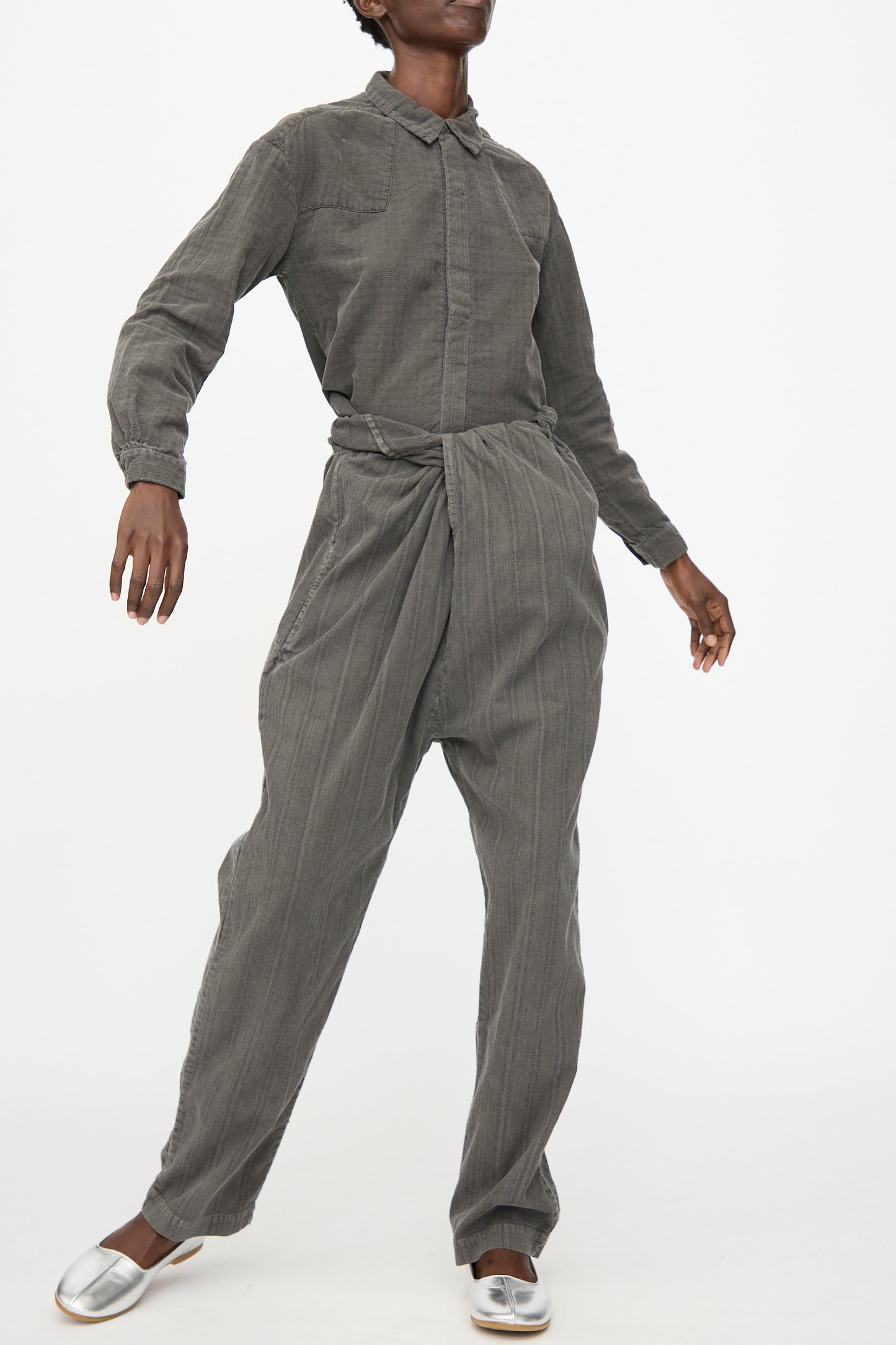 A person wears the Cosmic Wonder Stripe Combination Wrapped Pant in Dark Sumi, featuring a draped wrap detail, paired with a textured gray button-up shirt, standing against a plain white background.