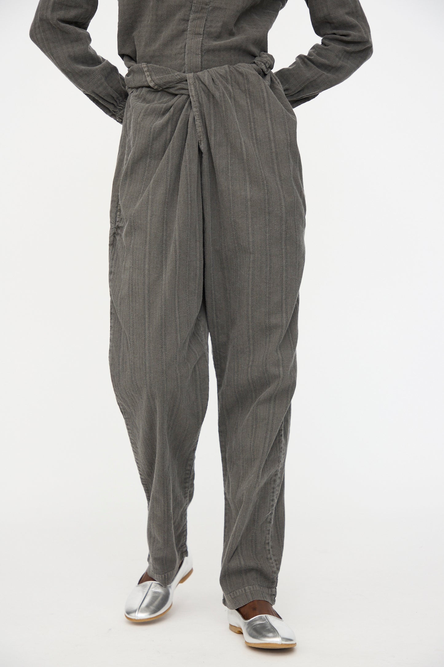 A person wears the Cosmic Wonder Stripe Combination Wrapped Pant in Dark Sumi—loose, textured gray organic cotton with a wrap waist—paired with silver slip-on shoes, standing against a plain white background.