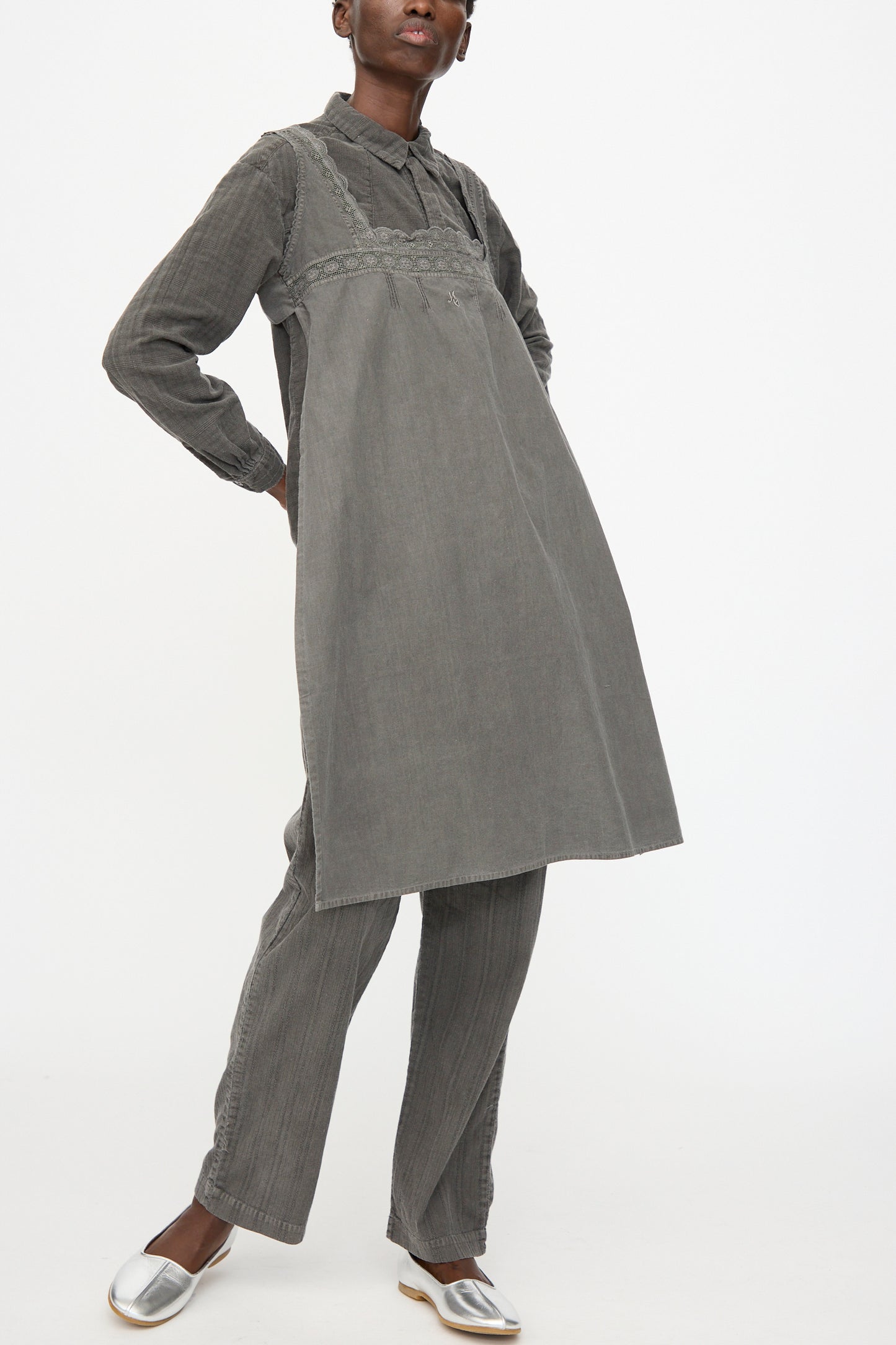 A person wears the Cosmic Wonder Taskuki Apron in Dark Sumi, styled over a gray long-sleeved shirt, matching gray pants, and silver shoes, standing against a plain white background.