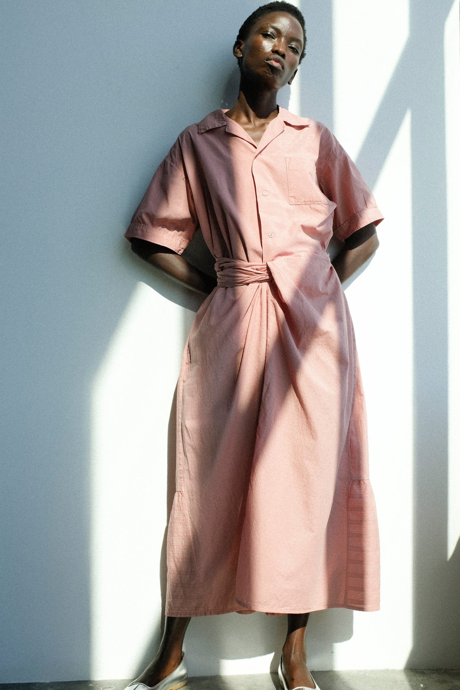 Woman wearing a pink dress standing against a white wall with shadows.