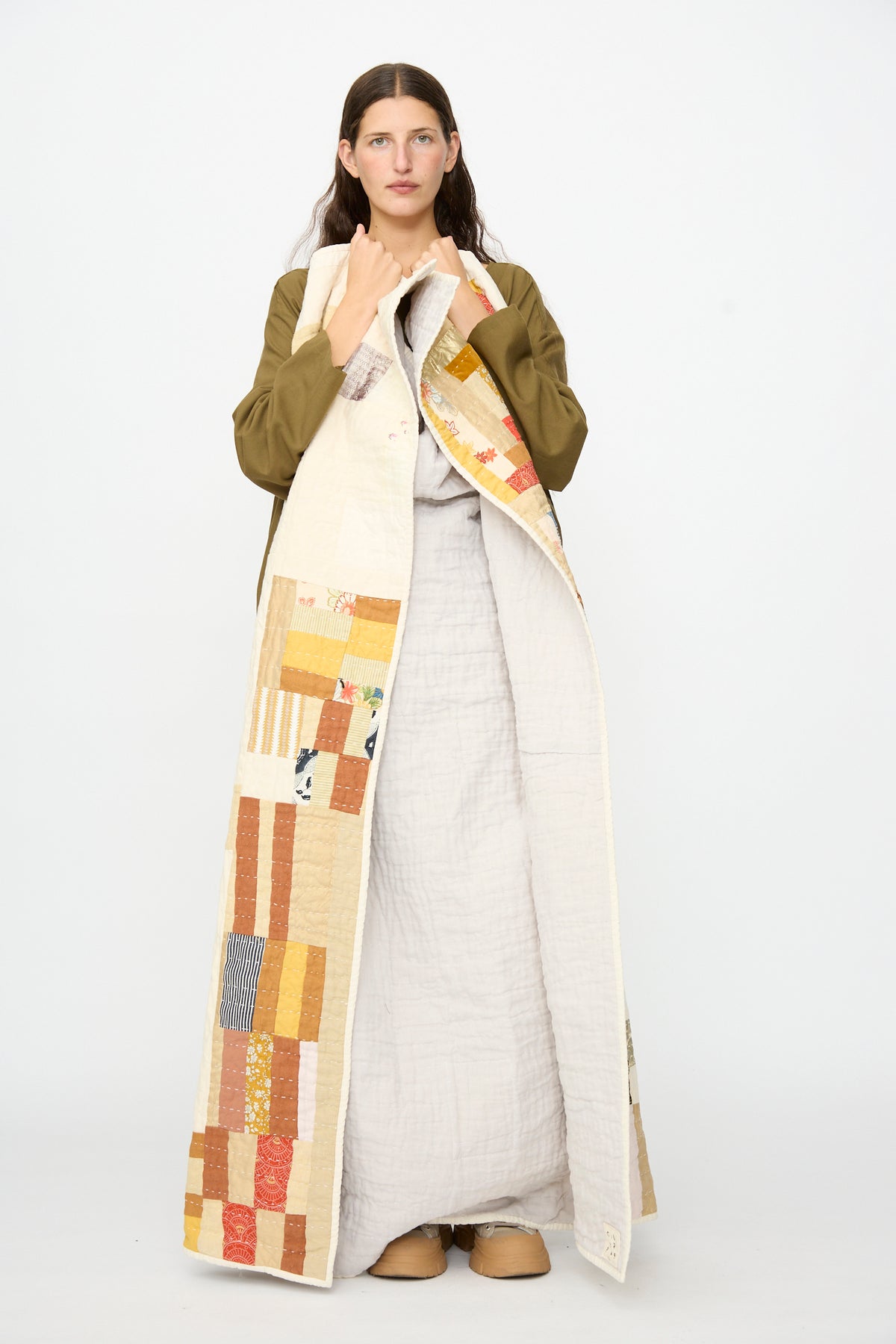 A person stands facing forward against a plain white background, wearing the Counterpane Patchwork Quilt in Yellow Gold Multi draped over a light dress and brown shoes.
