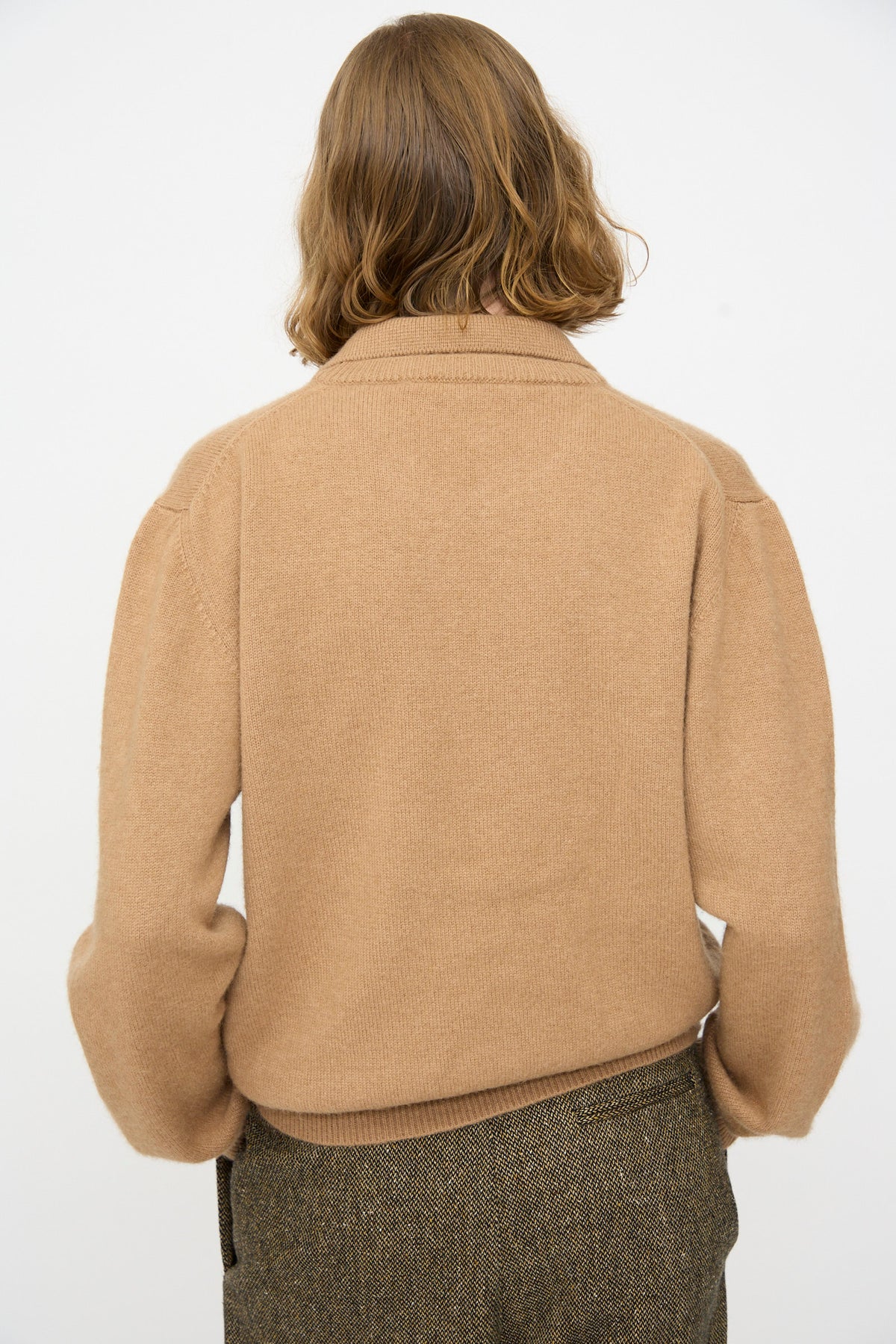 A person with medium-length light brown hair is seen from behind, wearing the Cristaseya Baby Camel Hair Polo sweater in tan and brown pants, photographed against a plain white background.