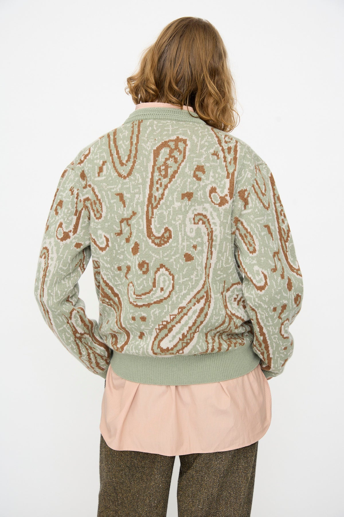 A person with shoulder-length light brown hair is seen from behind, wearing the Cristaseya Cashmere Jacquard Sweater in Sauge over a light pink shirt and paired with brown pants, set against a plain white background.