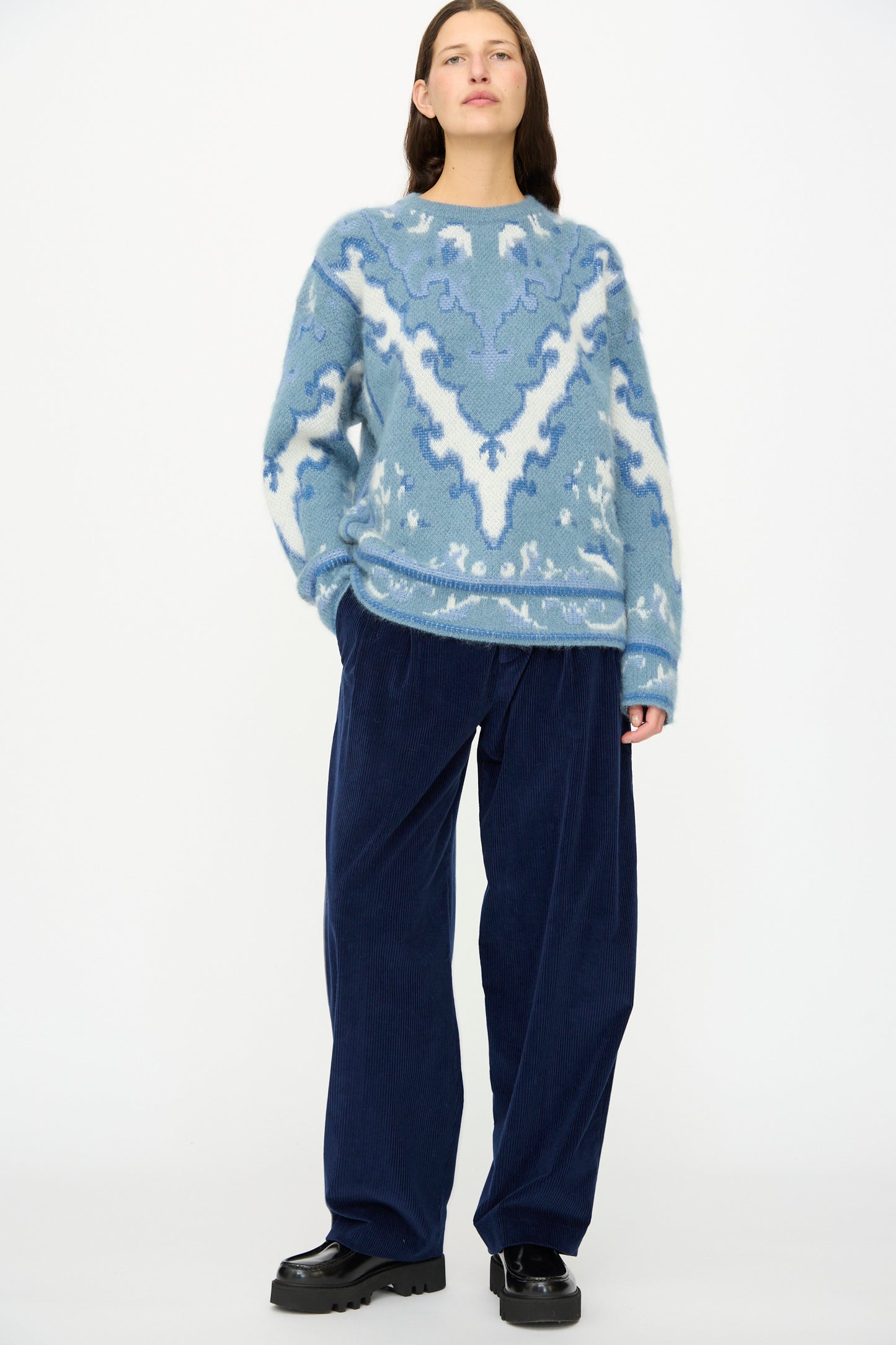 Person standing against a white background, wearing a blue patterned sweater, Cristaseya Corduroy Double Pleated Wide Pants in Indigo, and black shoes.