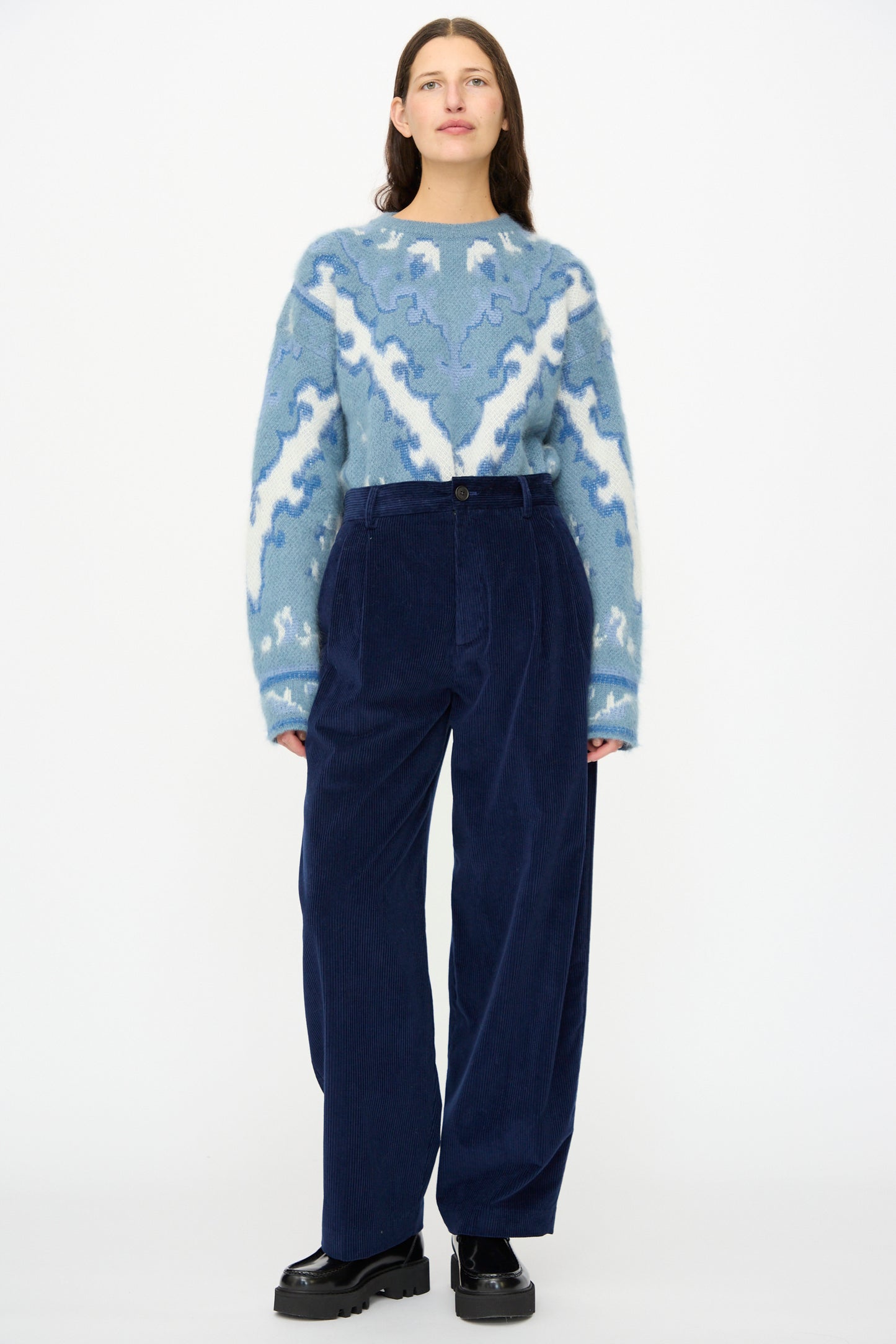 A woman stands against a white background wearing a blue patterned sweater, Cristaseya Corduroy Double Pleated Wide Pants in Indigo, and black shoes.