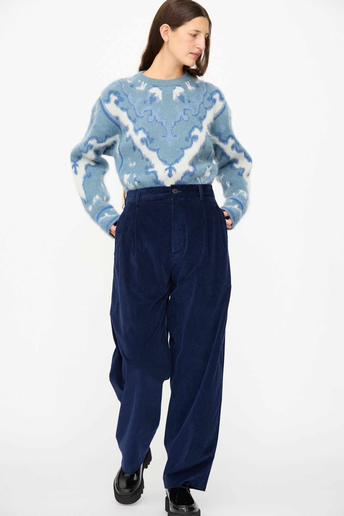 A woman stands with hands in pockets, wearing a patterned blue and white sweater, Cristaseya Corduroy Double Pleated Wide Pants in Indigo, and black shoes against a plain white background.