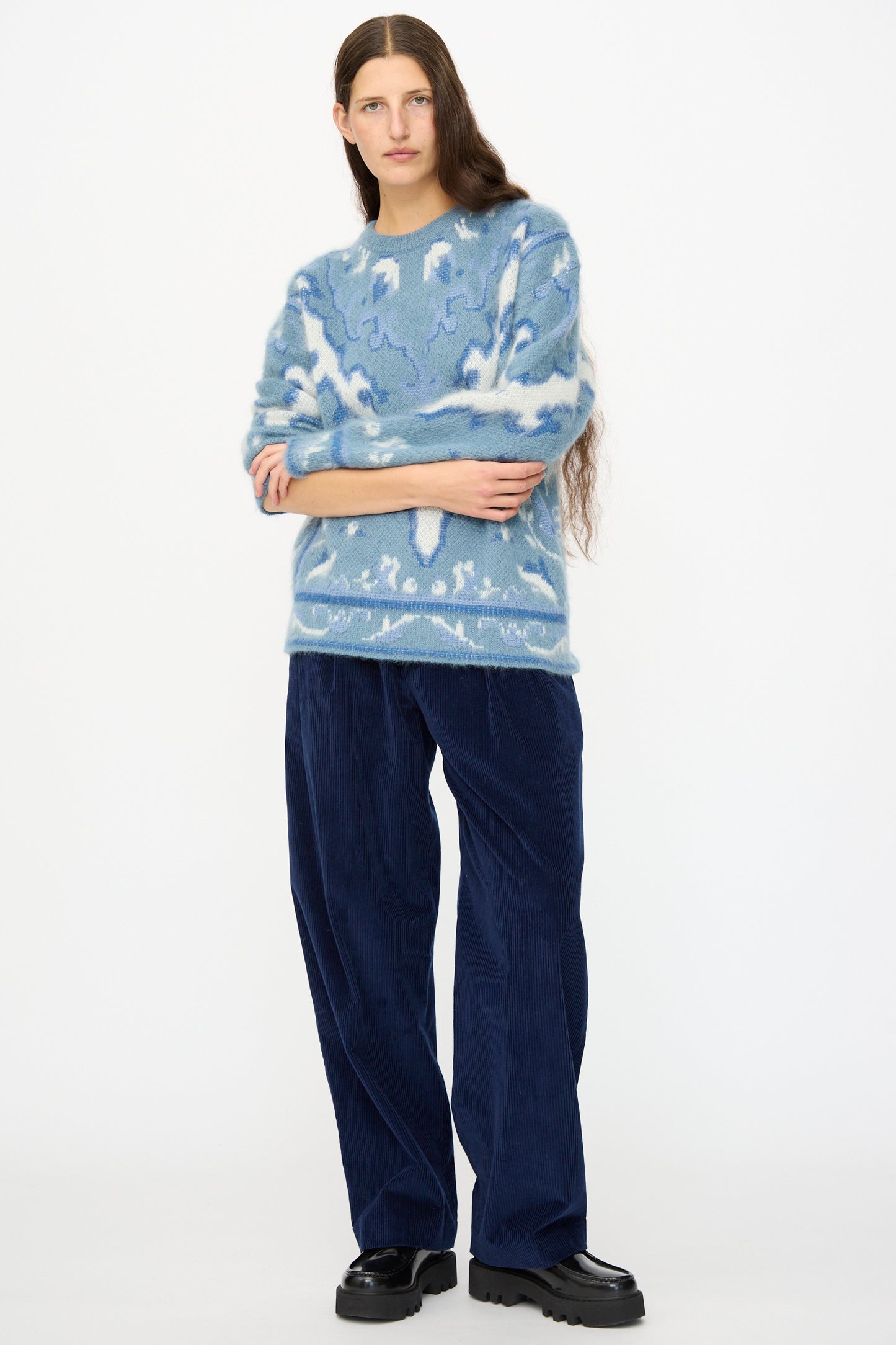 A person with long hair wears a blue patterned sweater, Cristaseya Corduroy Double Pleated Wide Pants in Indigo, and black platform shoes, standing against a plain white background.