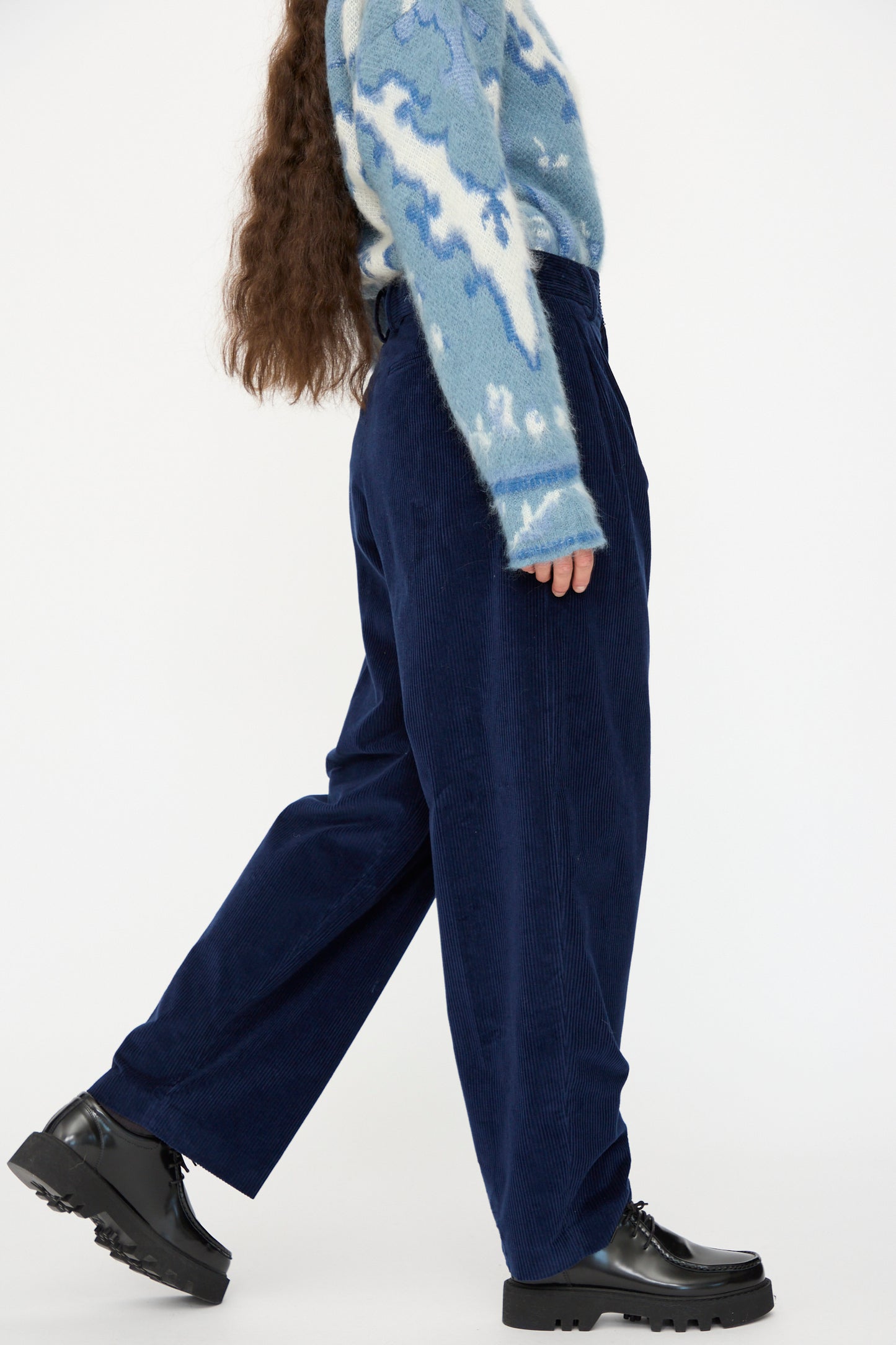 A person walks against a plain white background wearing a blue and white patterned sweater, Cristaseya Corduroy Double Pleated Wide Pants in Indigo, and black platform shoes.