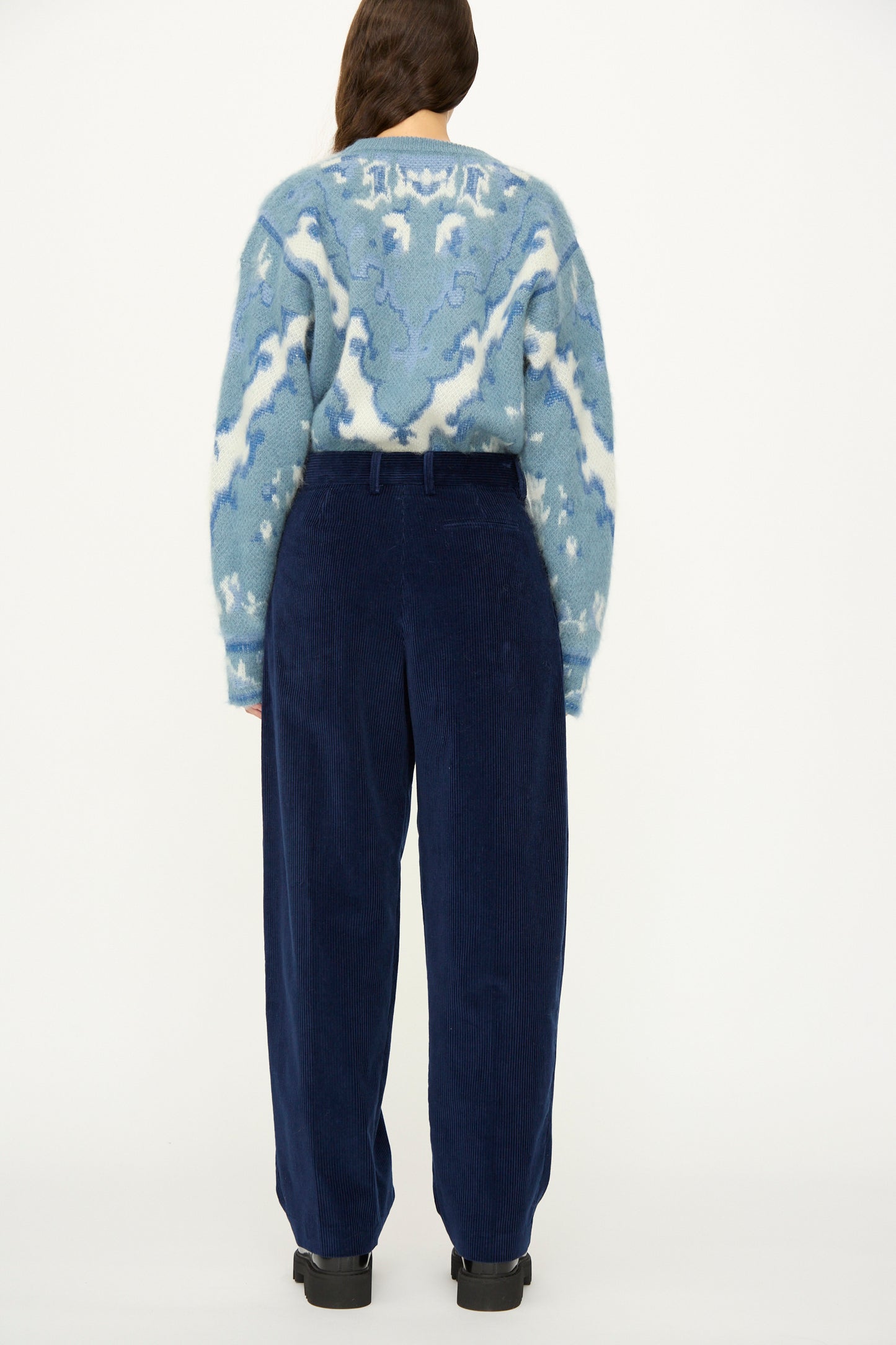 A person stands with their back to the camera wearing a blue and white patterned sweater, Cristaseya Corduroy Double Pleated Wide Pants in Indigo, and black shoes against a white background.