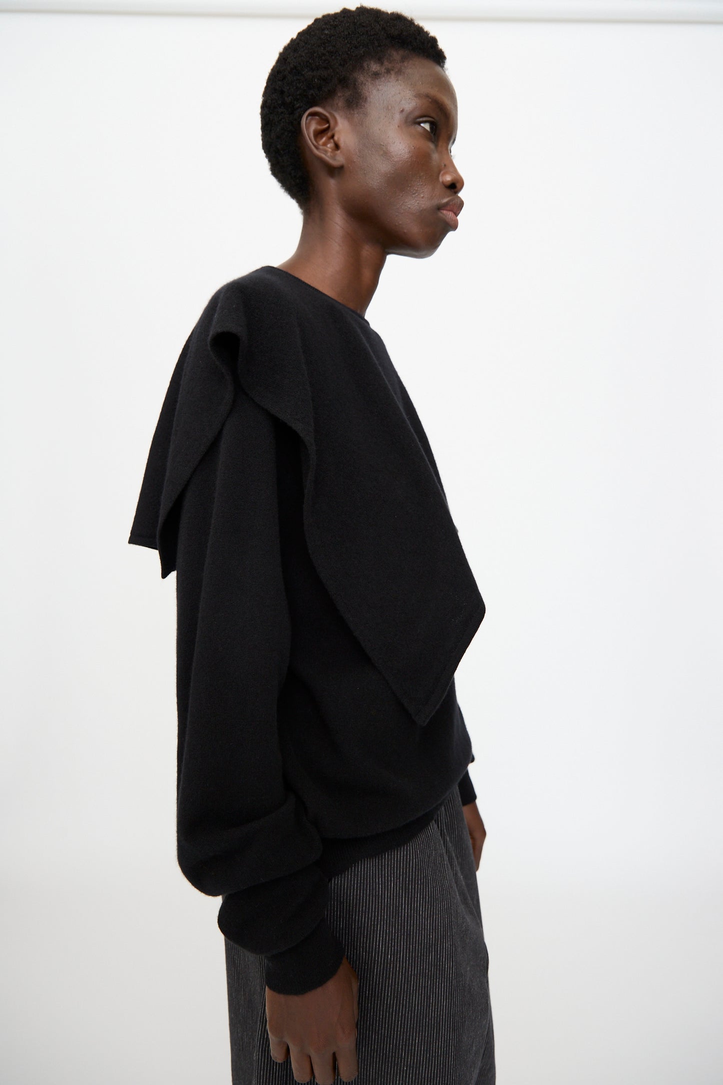 A person in profile stands against a plain background, wearing Cristaseya’s Flannel Sailor Collar Sweater in Black with extra black fabric over one shoulder, paired with striped pants.
