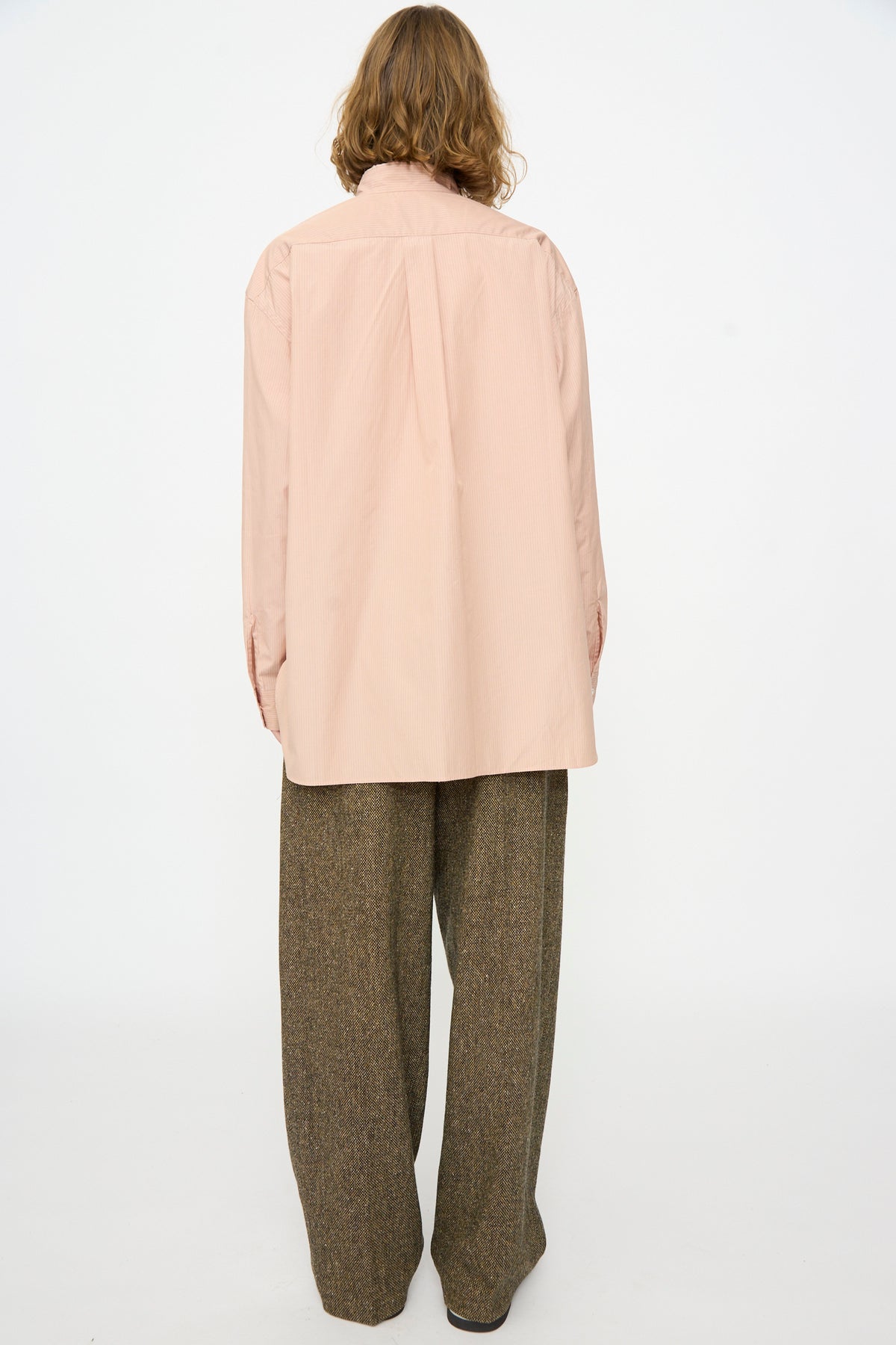 A person stands facing away from the camera against a plain white background, wearing the Cristaseya Handmade Fringed Mao Collar Shirt in pink and purple stripes with long sleeves, styled with wide brown trousers.