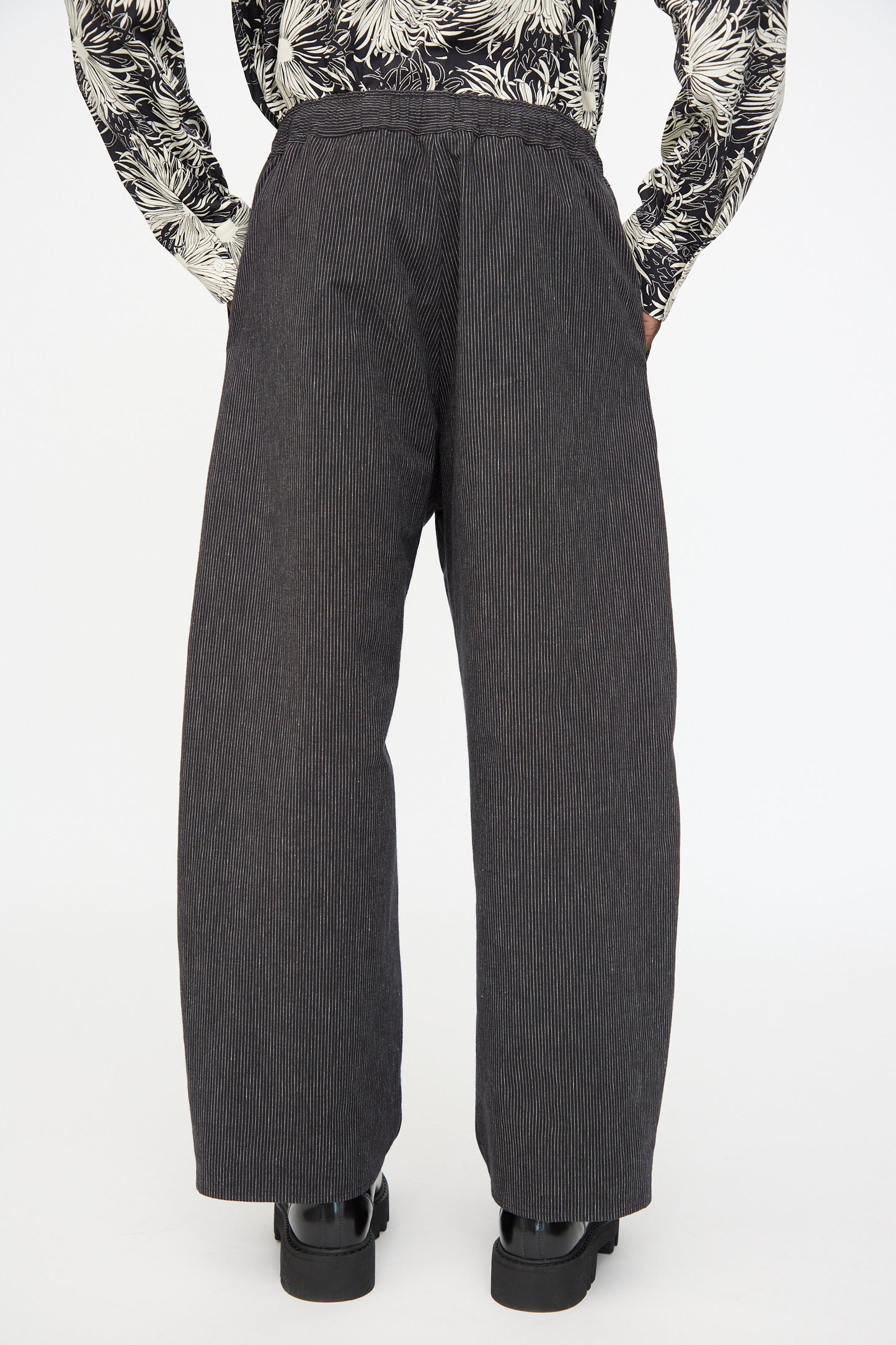 A person wearing Cristaseya Moroccan Pyjama Pants in Black with White Stripes, black shoes, and a patterned long-sleeve shirt, viewed from the back against a plain background.