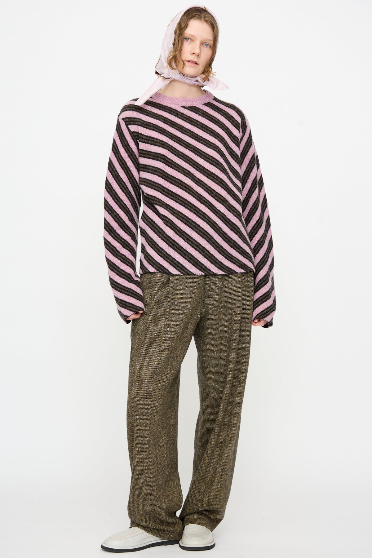 A person stands against a plain white background, dressed in a pink and black striped sweater, loose brown pants, white shoes, and the Cristaseya Silk Carré in Light Pink Checks worn as a headscarf.