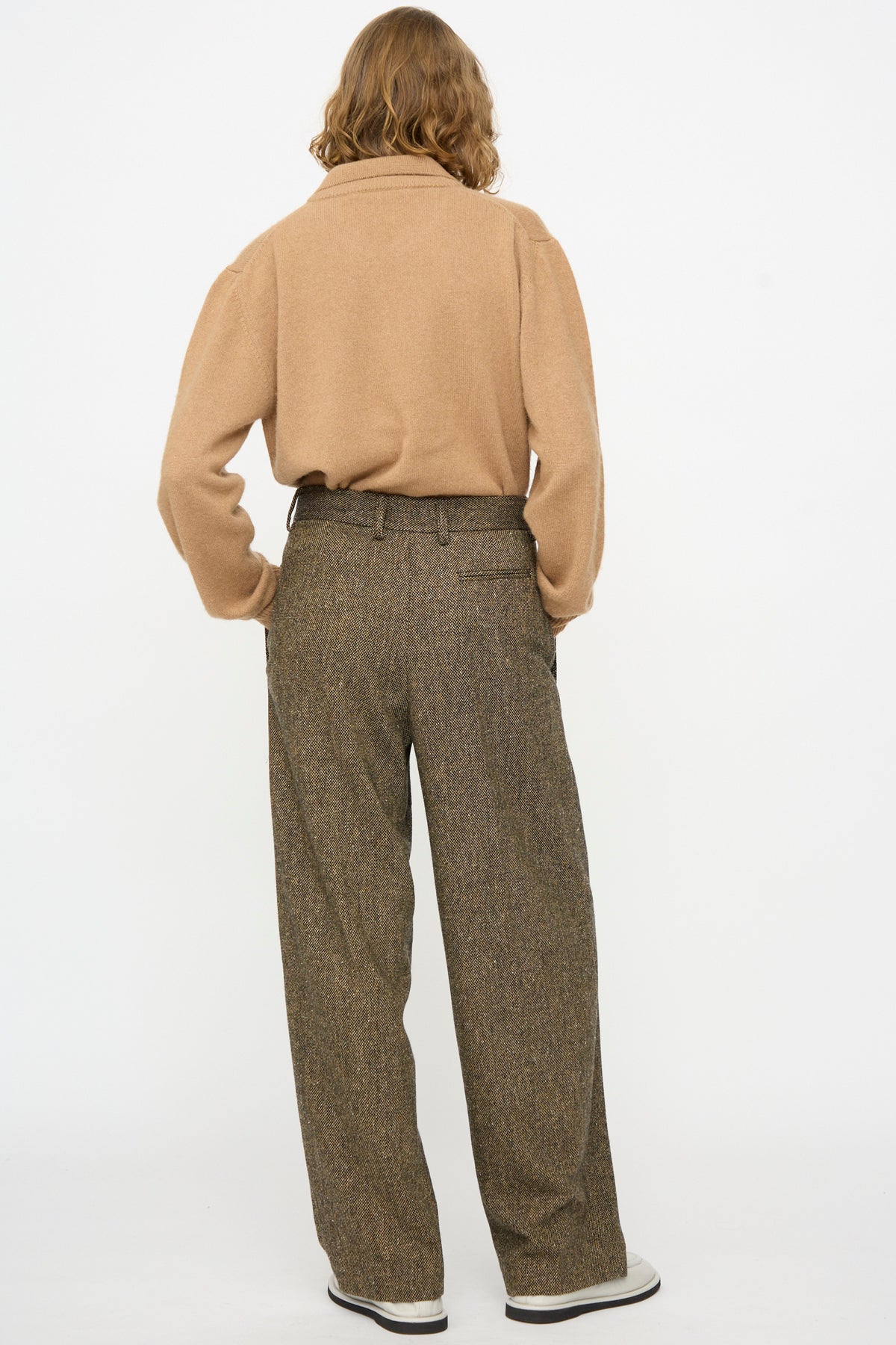 A person stands with their back to the camera, wearing a tan sweater and Cristaseya Tweed Double Pleated Wide Pants in Brown and Black, paired with white shoes, against a plain white background.