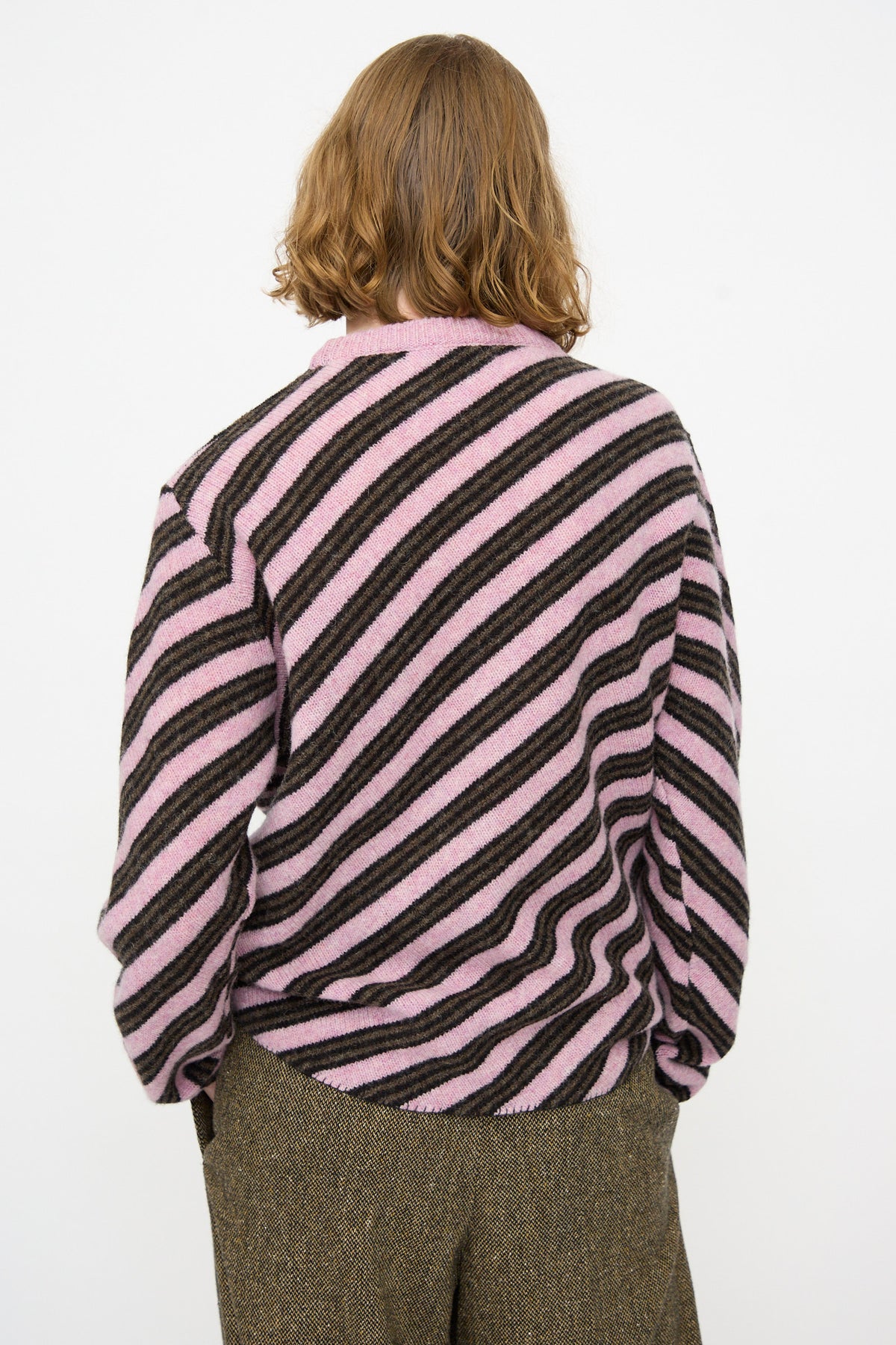 A person with shoulder-length hair faces away, wearing the Cristaseya Wool Diagonal Striped Sweater in Taupe and Pink and brown textured pants against a plain white background.