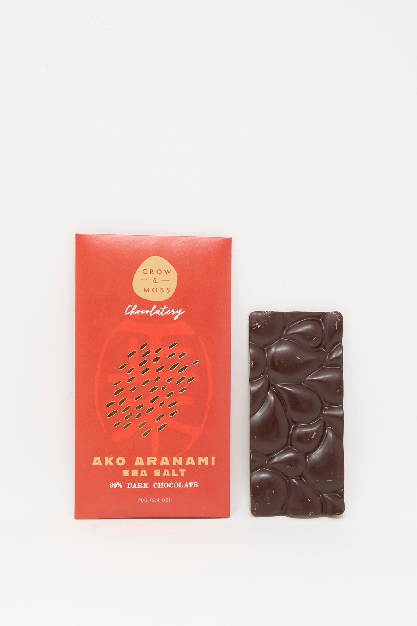 A red Crow & Moss Ako Aranami Sea Salt Dark Chocolate wrapper lies beside an unwrapped, textured dark chocolate bar, both displayed on a white background.