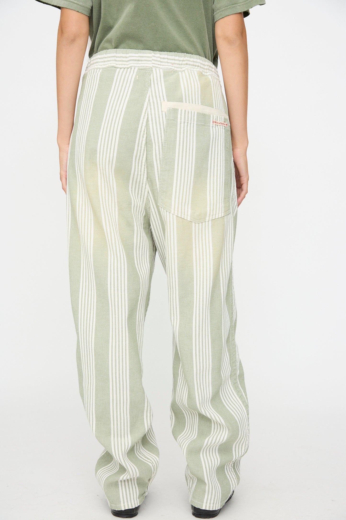 A person is seen from the back wearing Dr. Collectors’ 9 oz. P43 Kyoto Multi Stripe Pant in Sunfaded Light Green, which features loose, high-waisted relaxed cotton construction with vertical green and white stripes. The elastic waistband provides comfort, and a red label accents one of the back pockets.