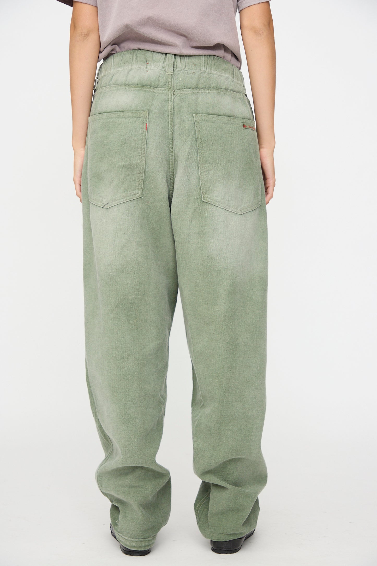 A person is seen from the back wearing Dr. Collectors’ P40 Z Boys Pant in Sunfaded Army Green—a relaxed fit with a loose silhouette, crafted from soft Japanese cotton. The pants feature two back pockets and a comfortable elastic waistband, set against a plain white background.