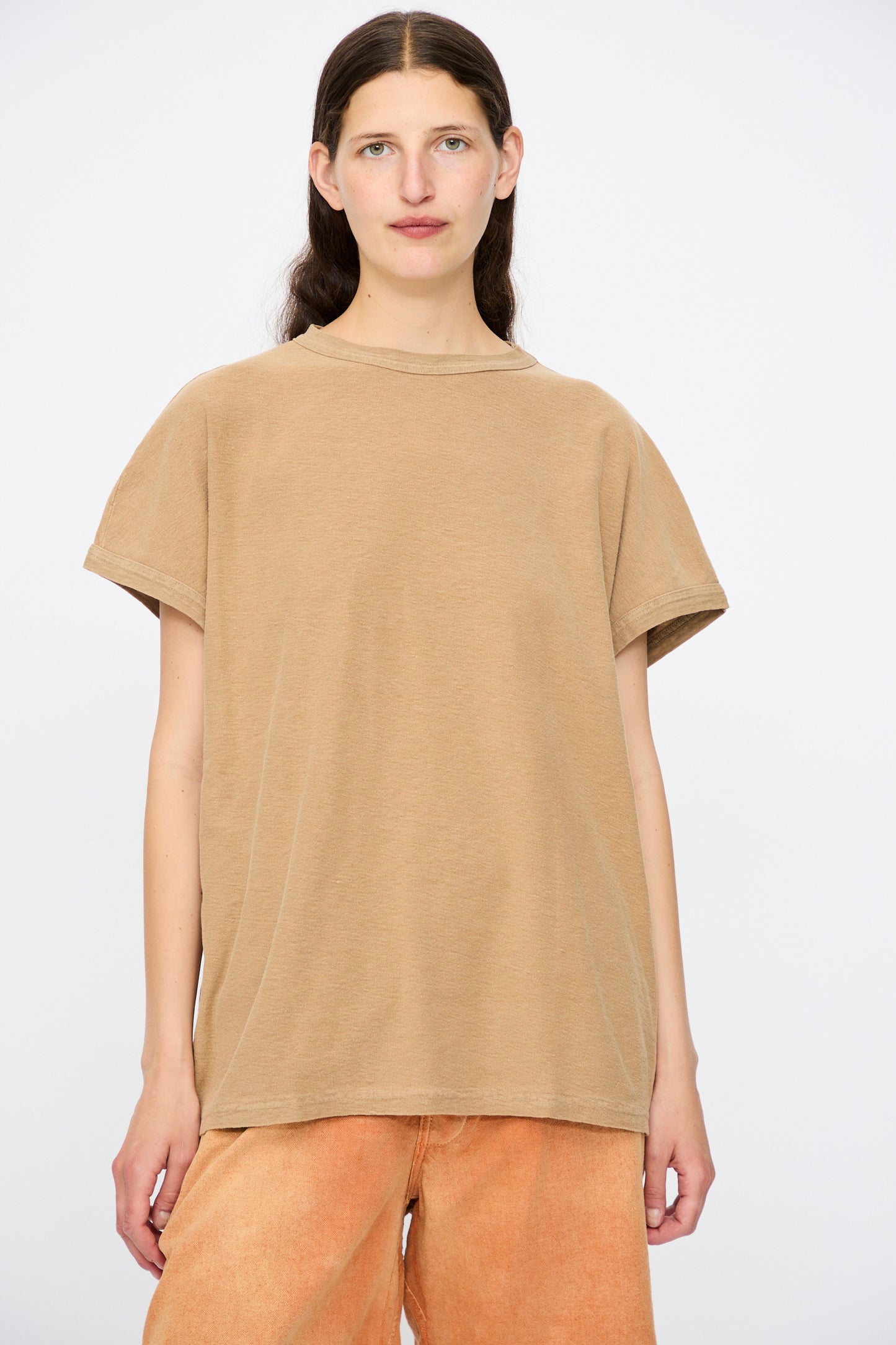 A person with long brown hair wears the Dr. Collectors Palm Springs Tee Sunfaded in Mud, styled with peach-colored pants, standing against a plain white background.