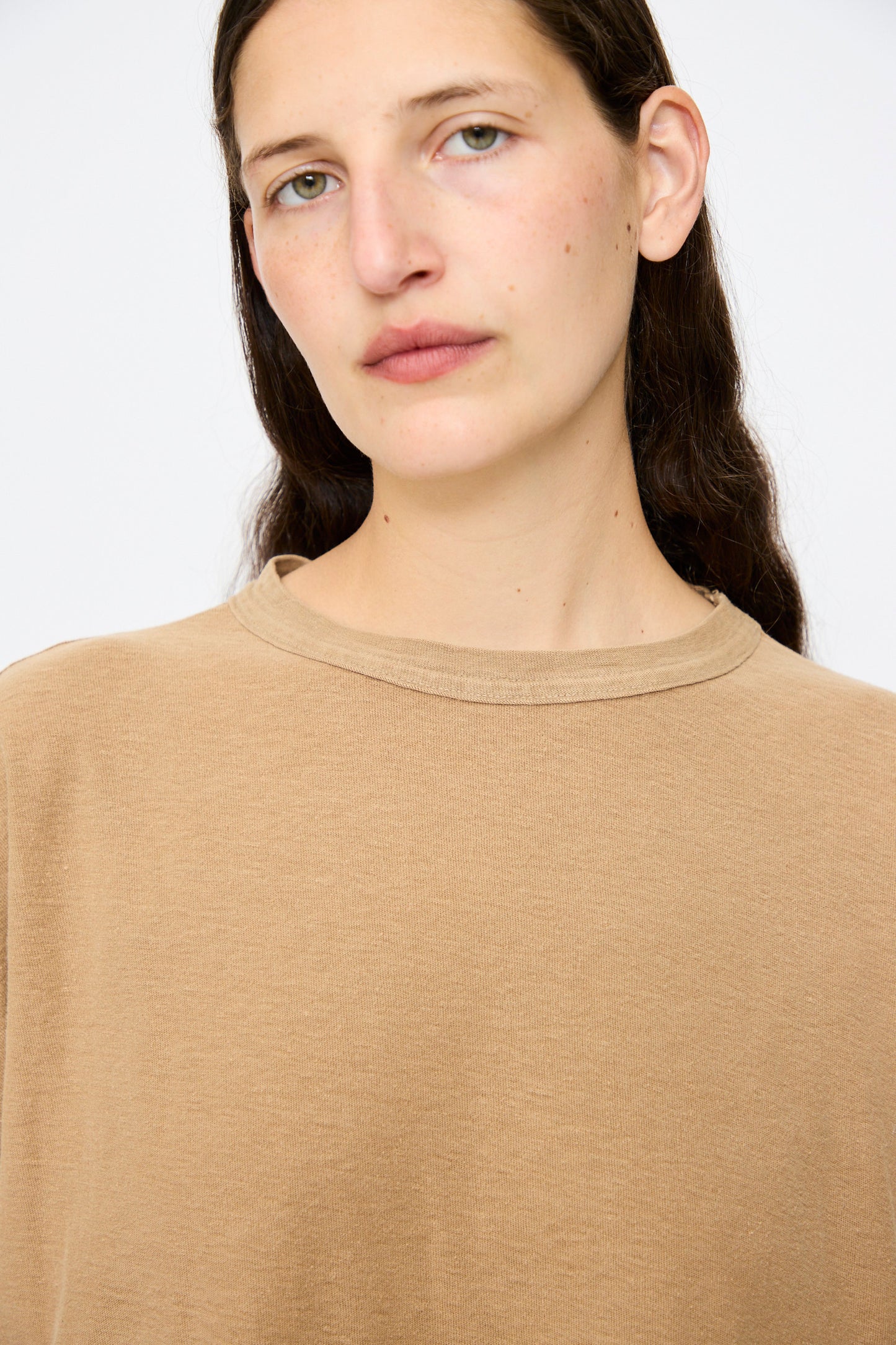 A person with long brown hair and light skin wears the Dr. Collectors Palm Springs Tee Sunfaded in Mud, made with organic cotton and plant-based dyes, standing against a plain white background.