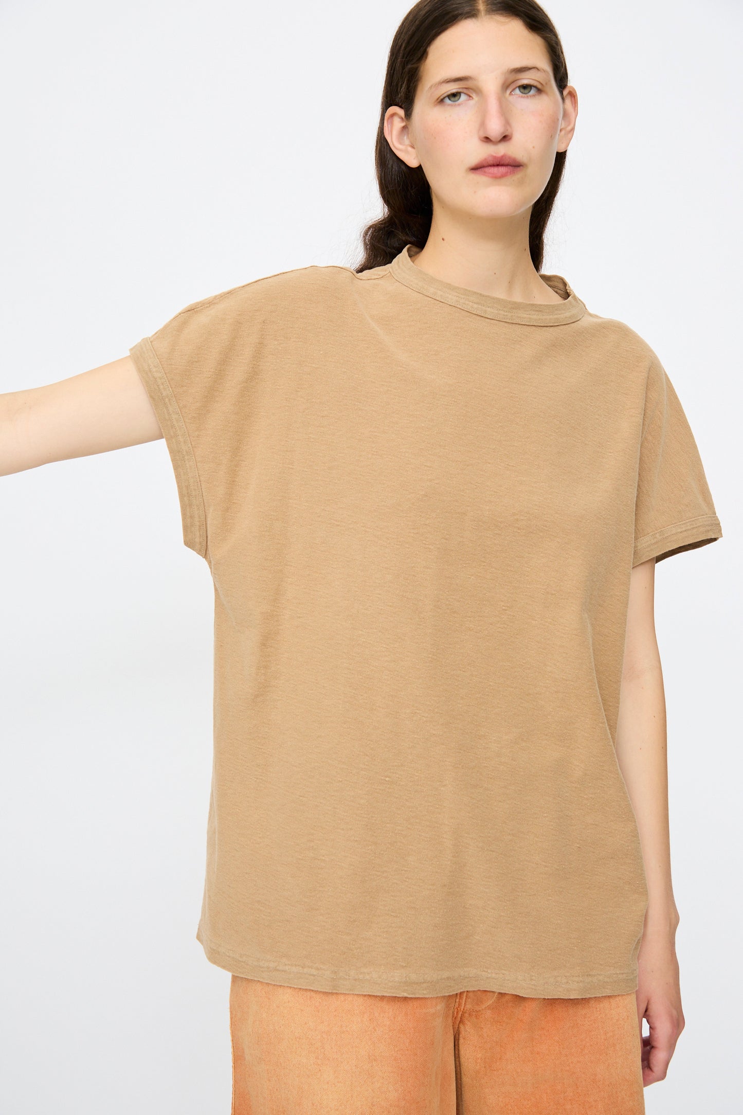 A person stands against a plain background wearing the Dr. Collectors Palm Springs Tee Sunfaded in Mud, a loose-fitting tan organic cotton t-shirt made in California, paired with light orange pants and one arm extended to the side.