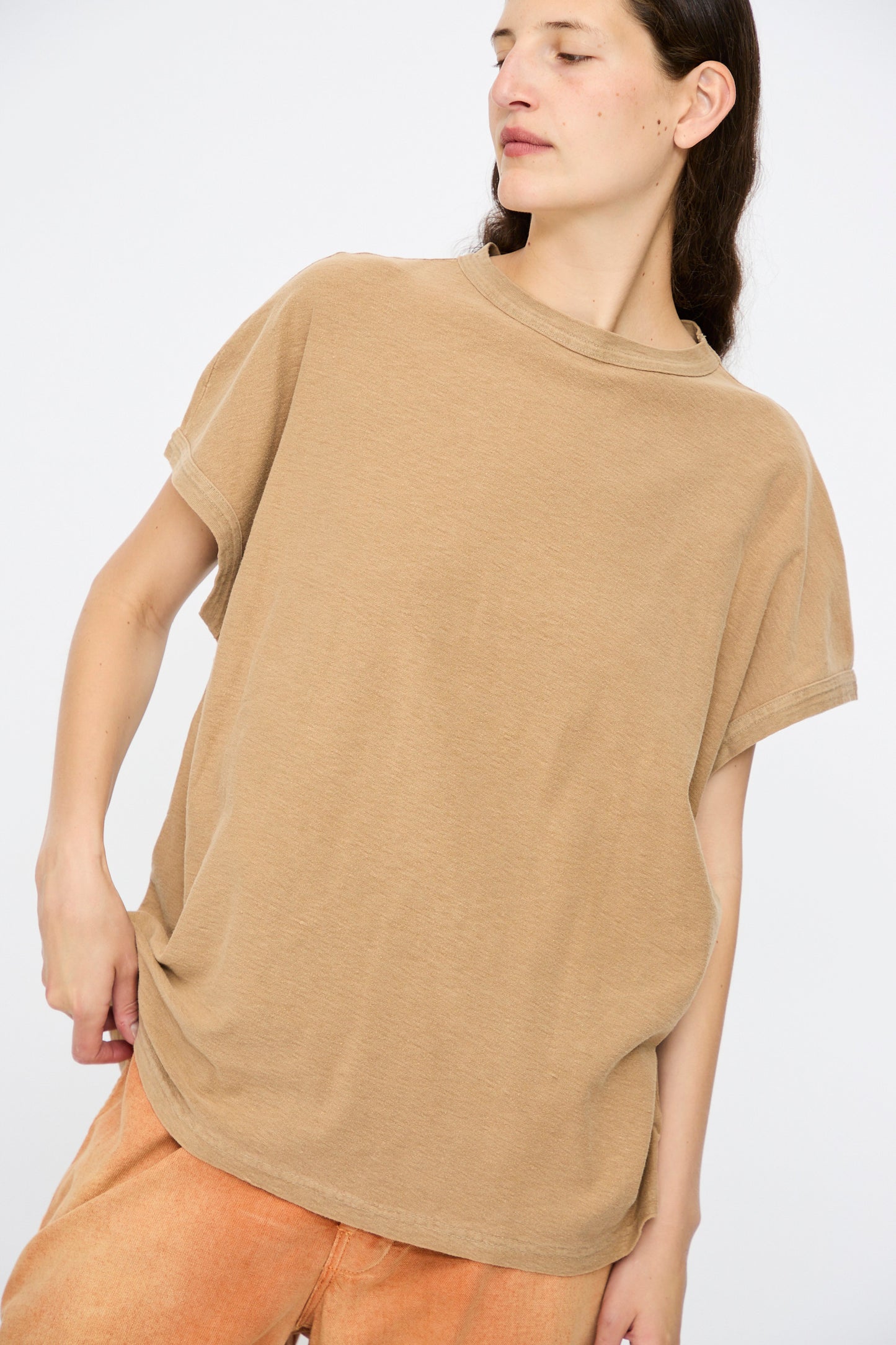 A person wears the Dr. Collectors Palm Springs Tee Sunfaded in Mud—an organic cotton, loose-fitting, short-sleeve top—paired with light orange pants against a plain light background.