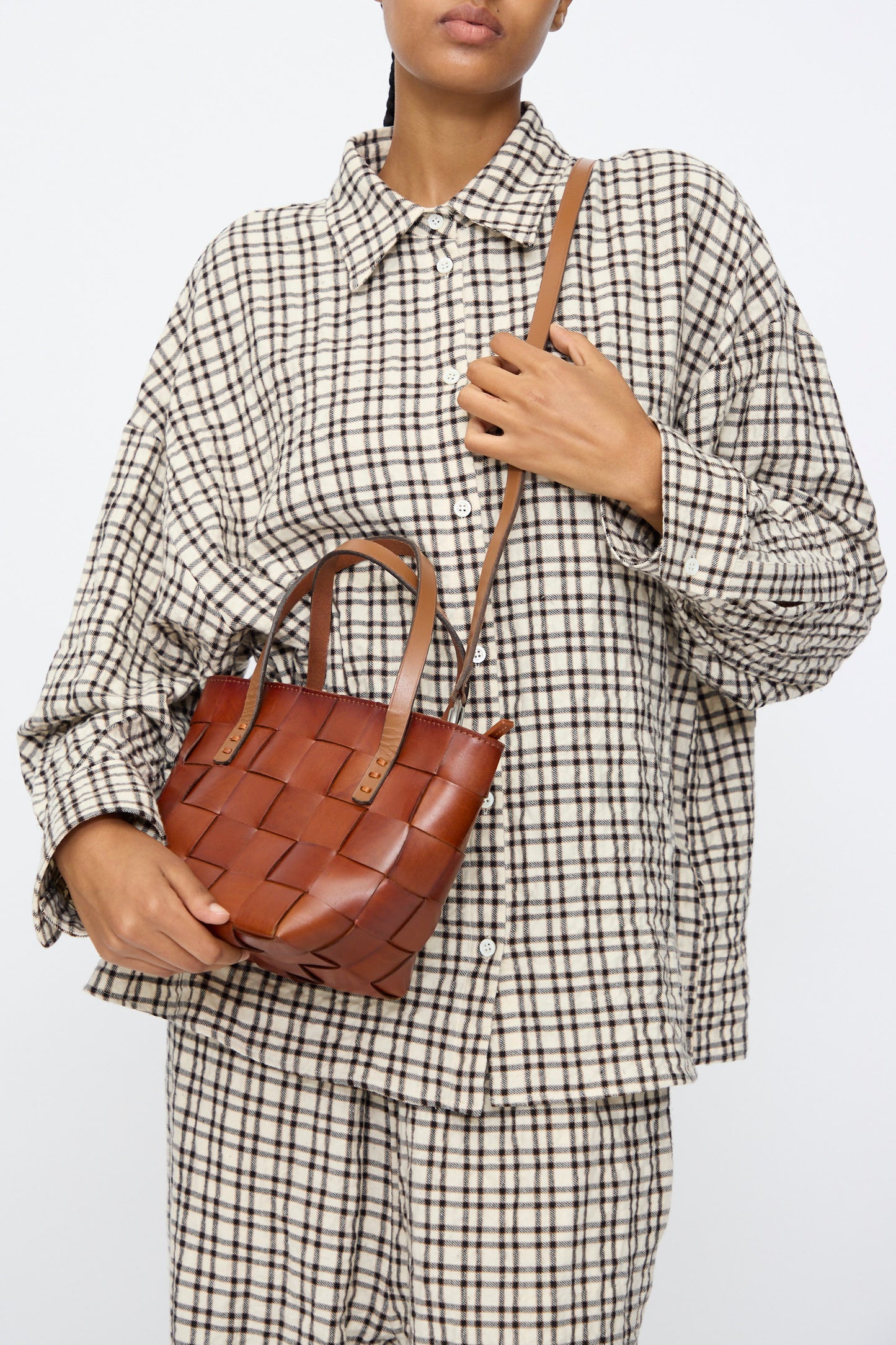 A person holds the Dragon Diffusion Japan Mini Tote in Tan, a small brown cow leather, basket-shaped bag with a shoulder strap, while wearing a loose checkered button-up shirt against a plain background.