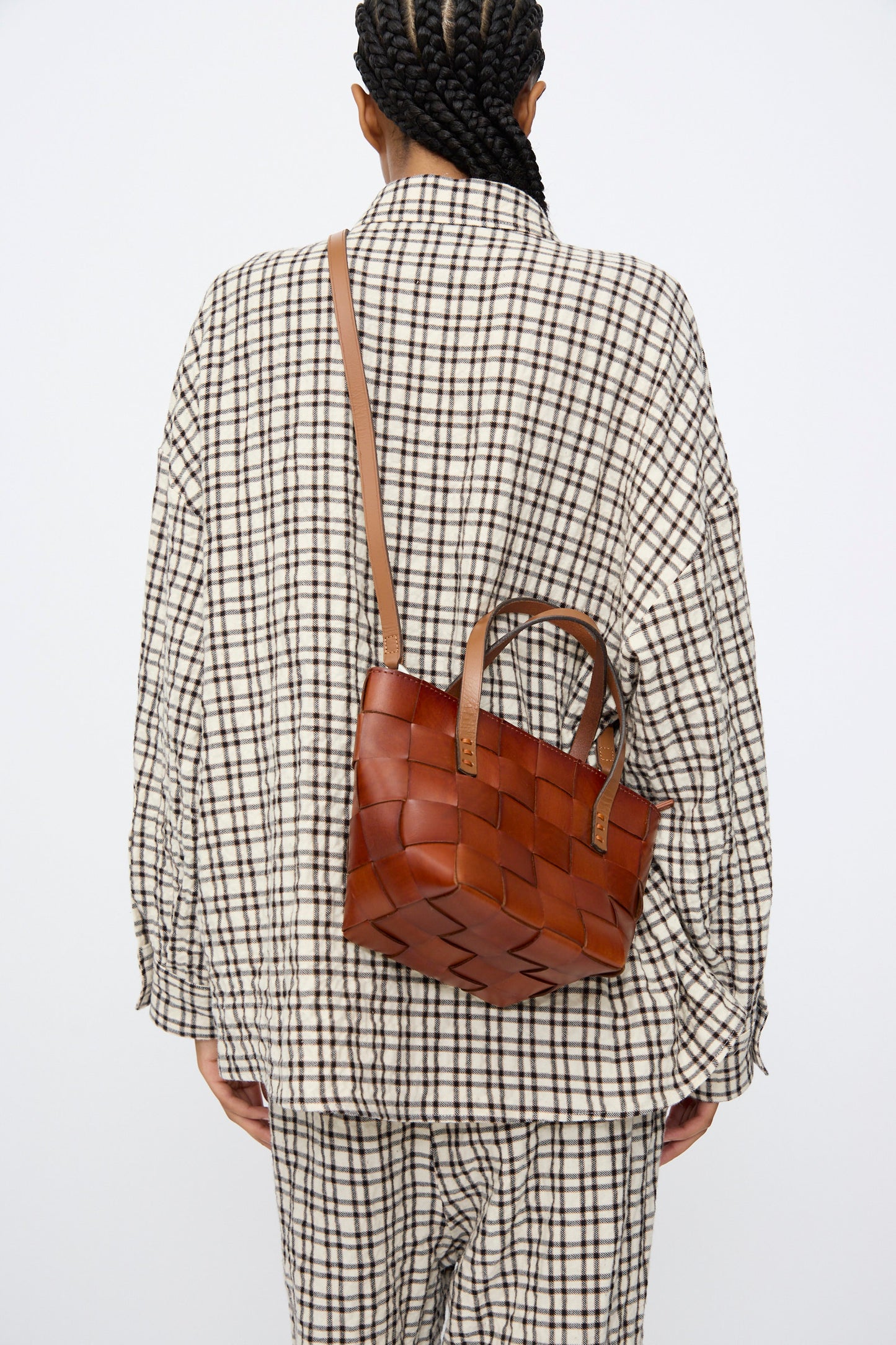 A person in a black and white checkered outfit, seen from behind, carries the Dragon Diffusion Japan Mini Tote in Tan—a brown cow leather, basket-shaped mini bag—over their shoulder.