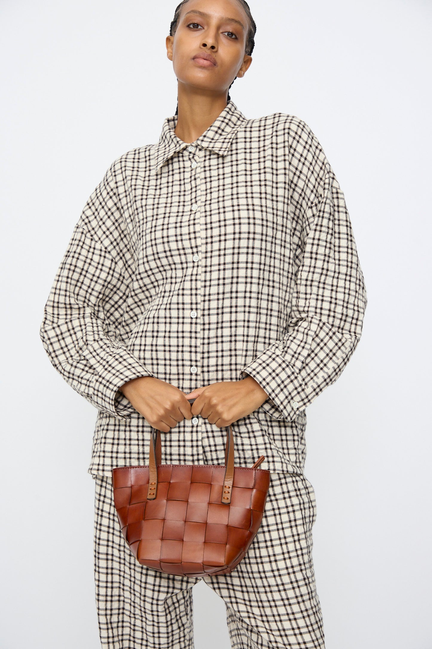 A person in a loose plaid shirt and pants set holds the Dragon Diffusion Japan Mini Tote in Tan, a small brown cow leather basket-shaped bag, against a plain white background.