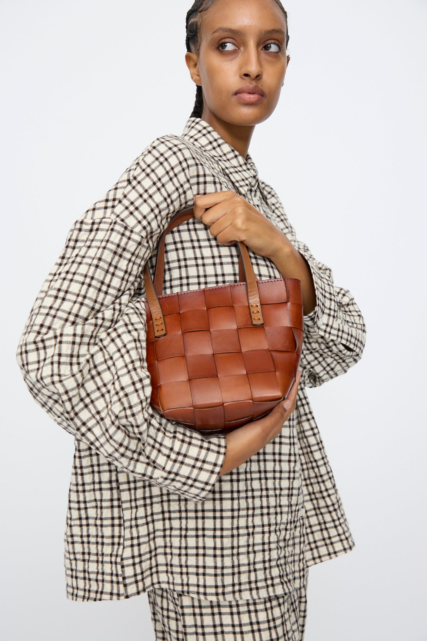 A person in a plaid shirt and matching pants holds the Dragon Diffusion Japan Mini Tote in Tan—crafted from woven cow leather—against a plain white background.