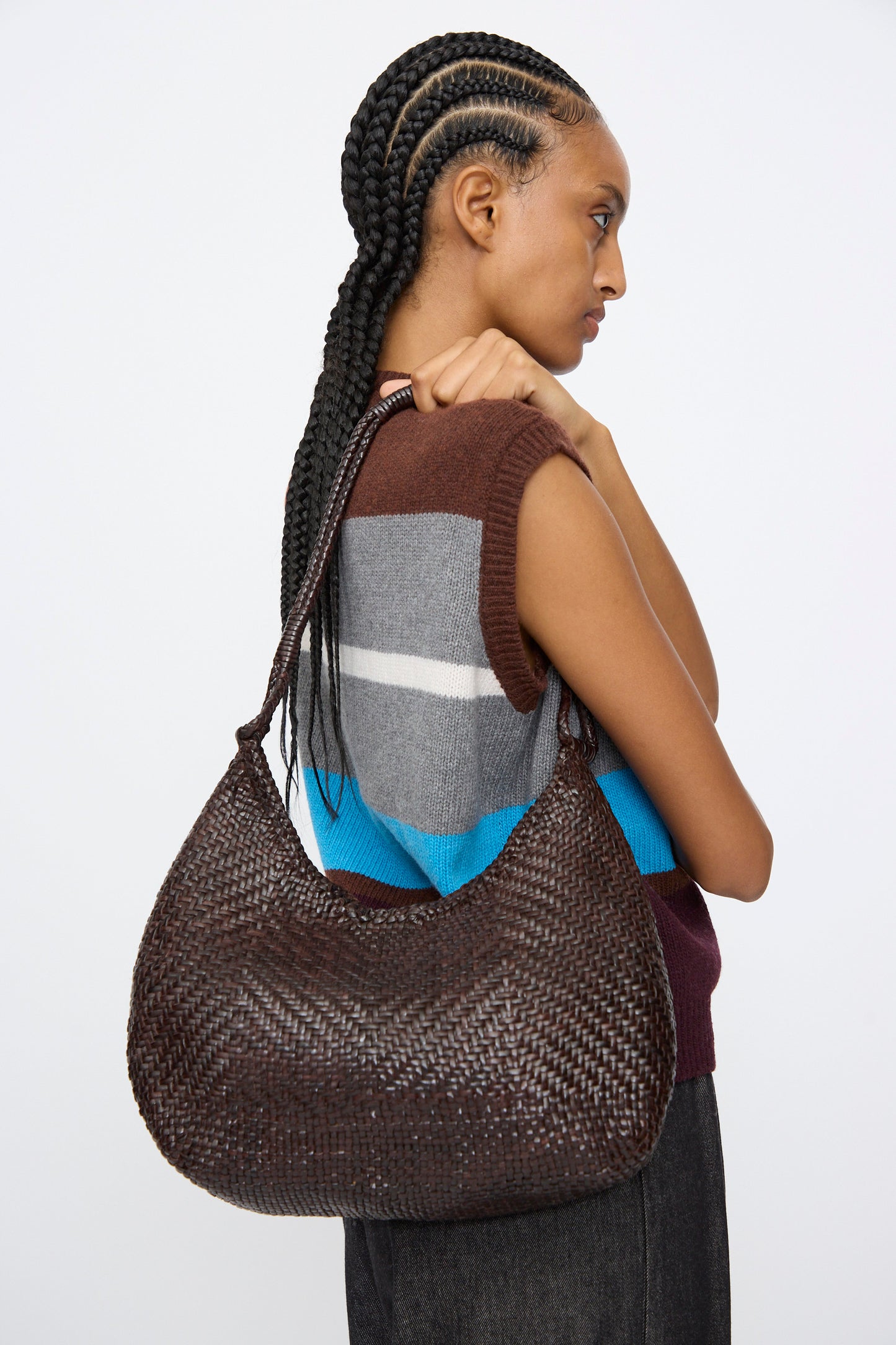 A person with braided hair in a sleeveless sweater holds the Dragon Diffusion Minga Hobo in Dark Brown, a large handwoven calf leather shoulder bag, over their shoulder against a plain background.