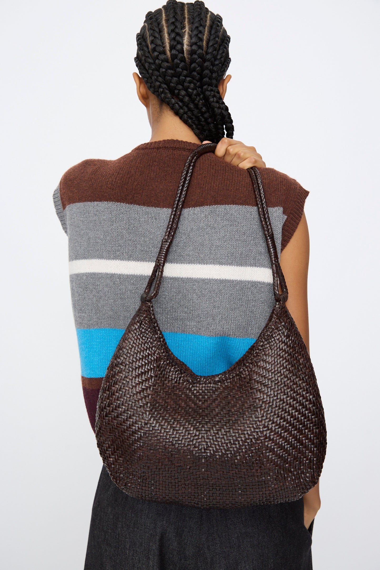 A person with braided hair is seen from the back wearing a sleeveless striped sweater and carrying Dragon Diffusion's Minga Hobo in dark brown, a large shoulder bag crafted from handwoven calf leather.