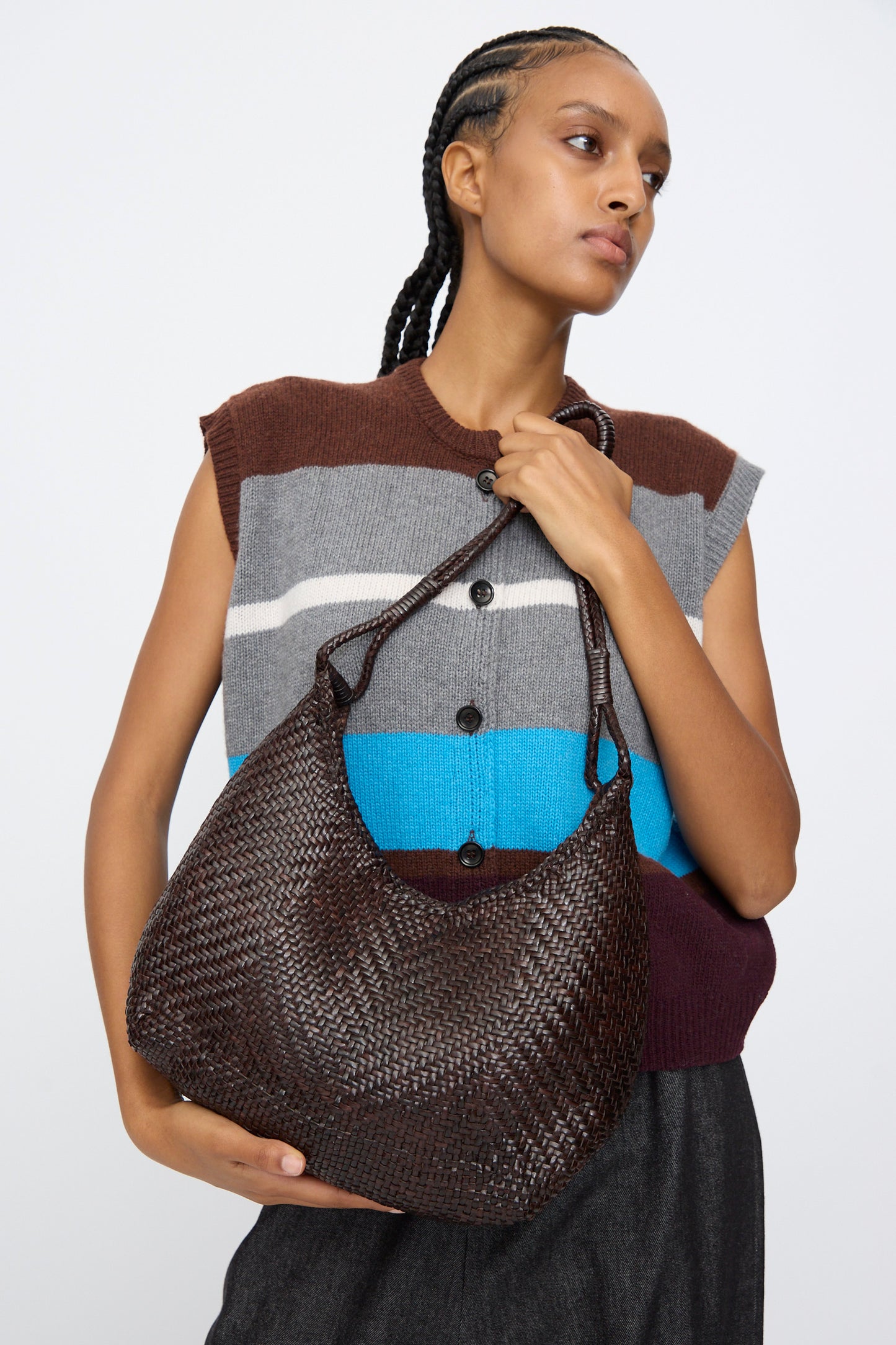 A person wearing a sleeveless, striped sweater and dark pants holds the Dragon Diffusion Minga Hobo in Dark Brown, a large handwoven calf leather shoulder bag, against a plain background.