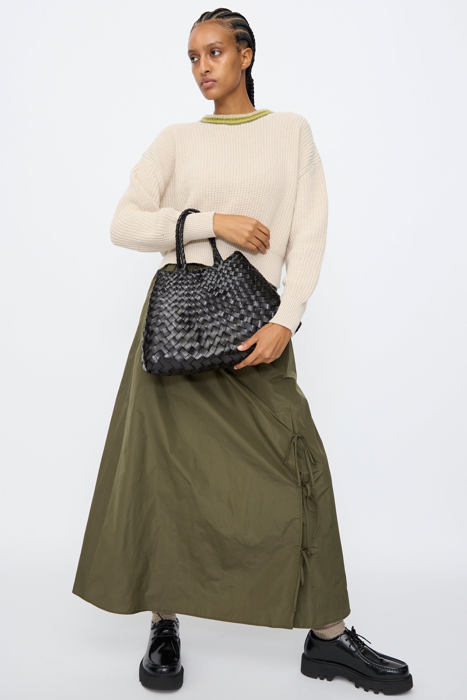 A person stands against a plain background wearing a beige sweater, olive green maxi skirt, black platform shoes, and holding the Dragon Diffusion Santa Croce Small bag in black with braided handles.