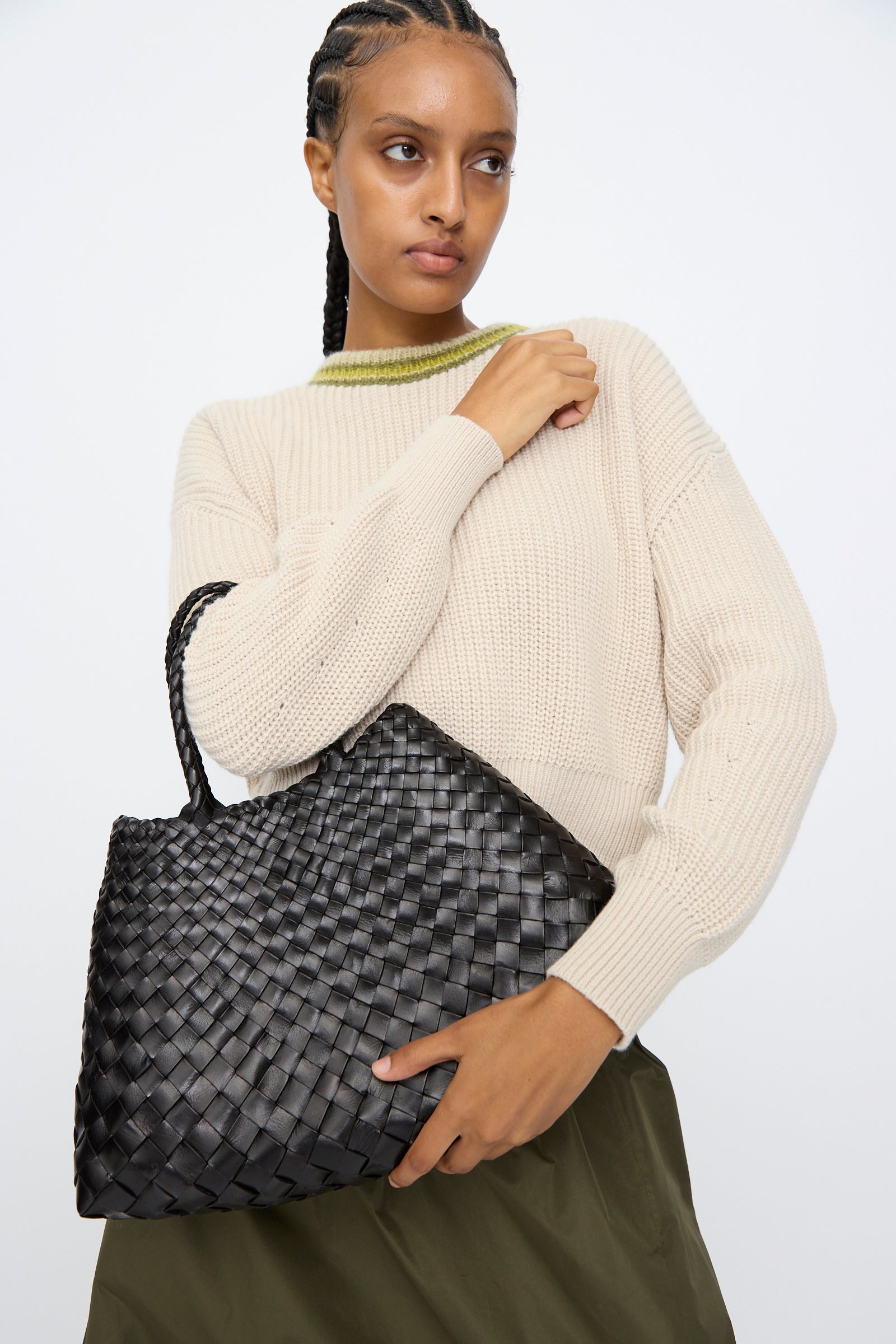 A woman in a beige sweater and olive skirt holds the Dragon Diffusion Santa Croce Small bag in black with braided handles against a plain white background.