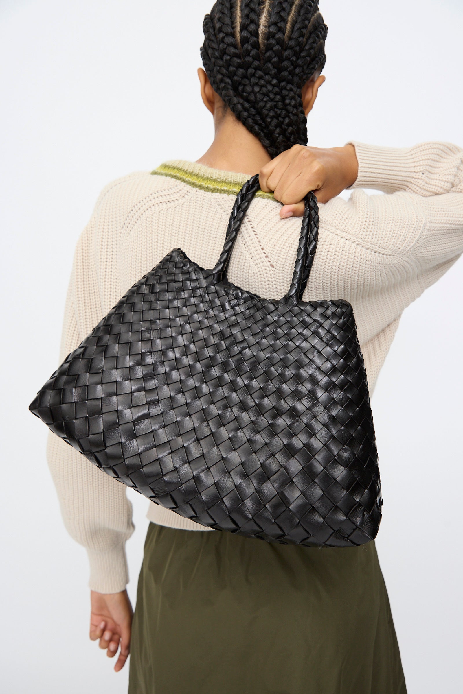 A person with braided hair carries the Dragon Diffusion Santa Croce Small in Black, a handwoven bag with braided handles, over their shoulder while wearing a cream sweater and olive green skirt.