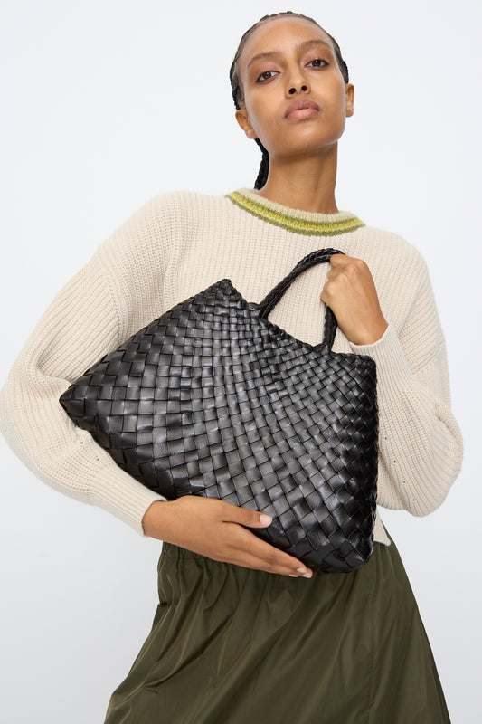 A person in a beige sweater and olive skirt holds the Dragon Diffusion Santa Croce Small bag in Black with braided handles against a plain background.