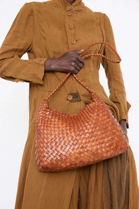 A person in a brown button-up dress holds a Dragon Diffusion Santa Rosa in Tan handwoven brown leather shoulder bag against a plain background.