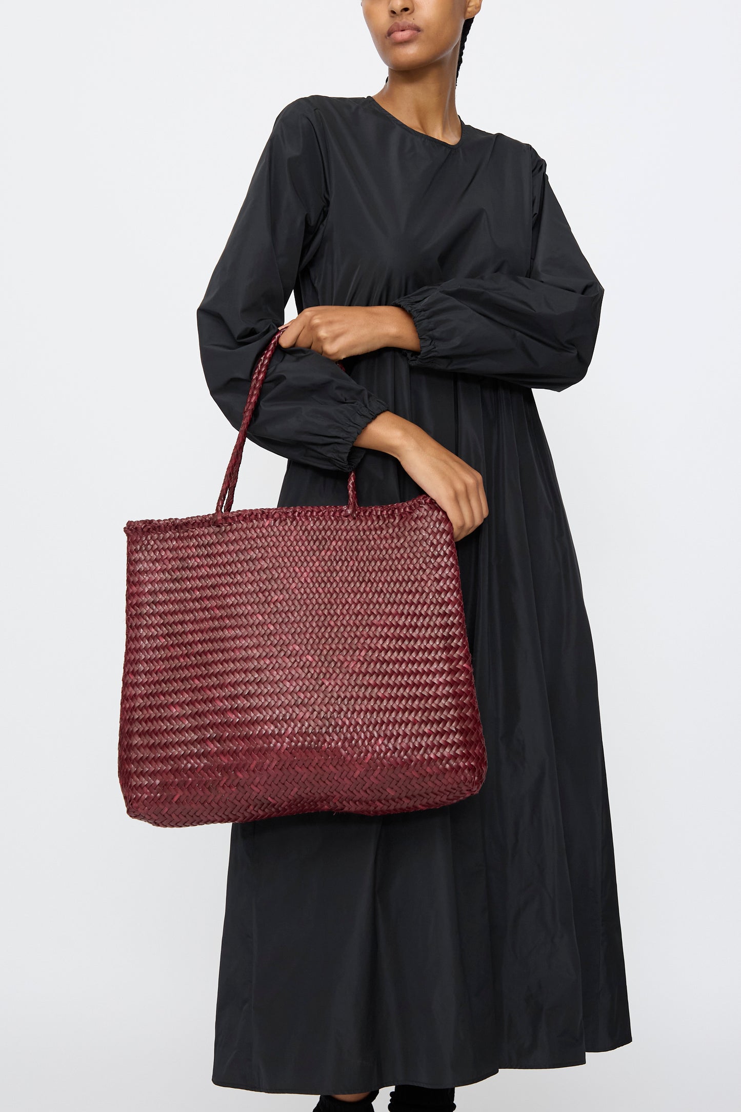 A person in a long-sleeved black dress holds the Dragon Diffusion Sophie Large bag in Bordo, a spacious handwoven leather accessory, against a plain white background.