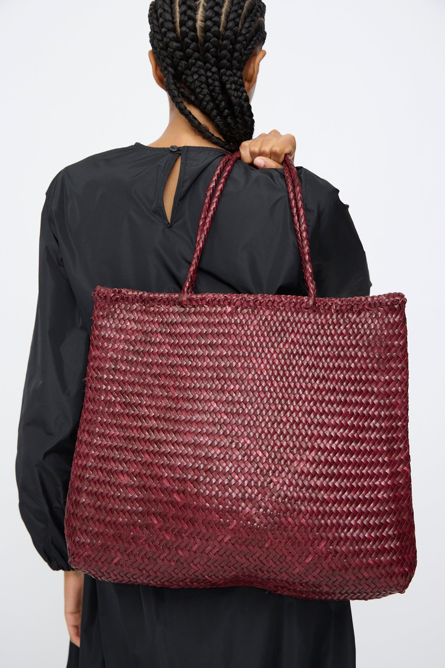 A person with braided hair, wearing a black top, carries the Dragon Diffusion Sophie Large in Bordo handwoven leather bag over their shoulder, facing away from the camera.